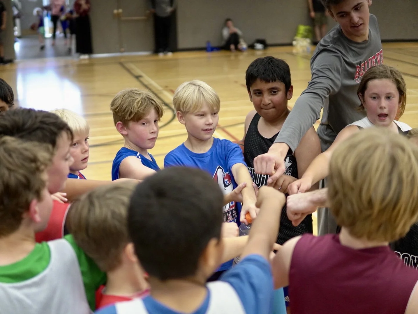 Young children in sports jerseys gather in a gymnasium, participating in a team-building activity with an adult coach.
