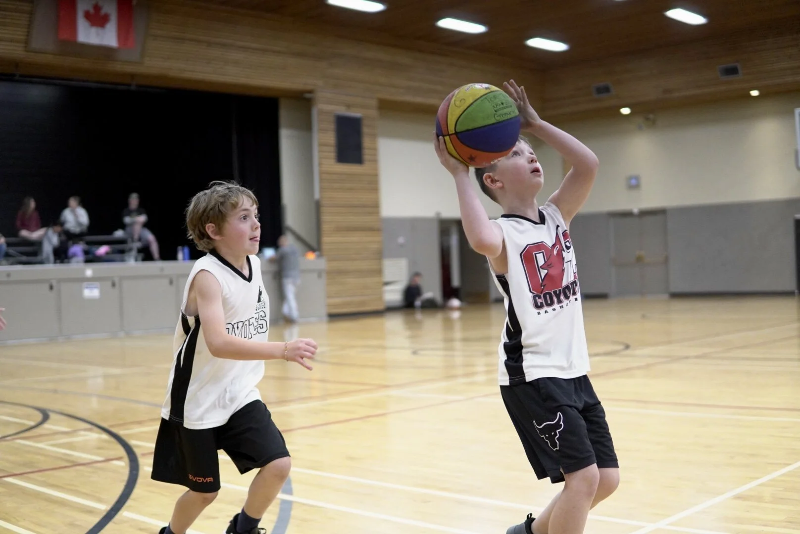Two young boys playing basketball in an indoor gymnasium; one is preparing to shoot with a colorful basketball, while the other is watching.