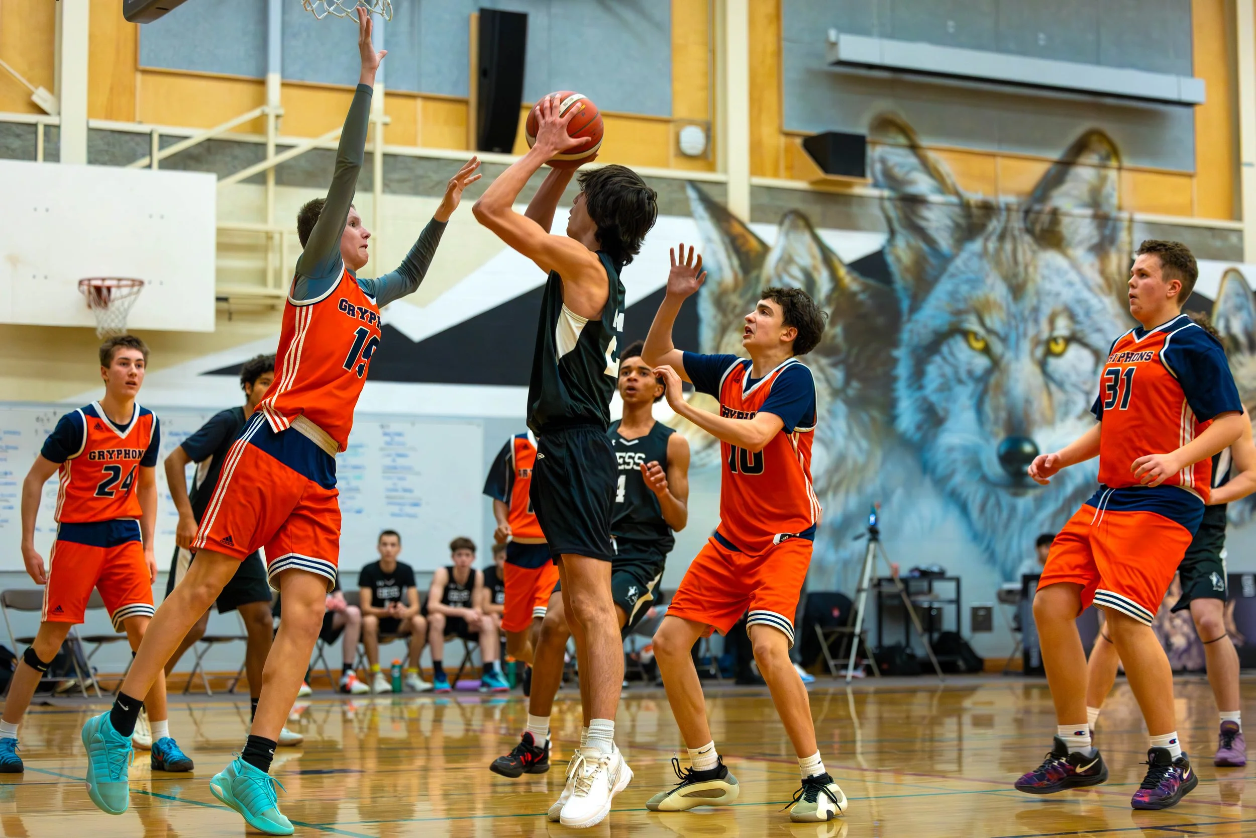 Young basketball players in a gym during a game, with one player attempting a shot while others try to block or defend.
