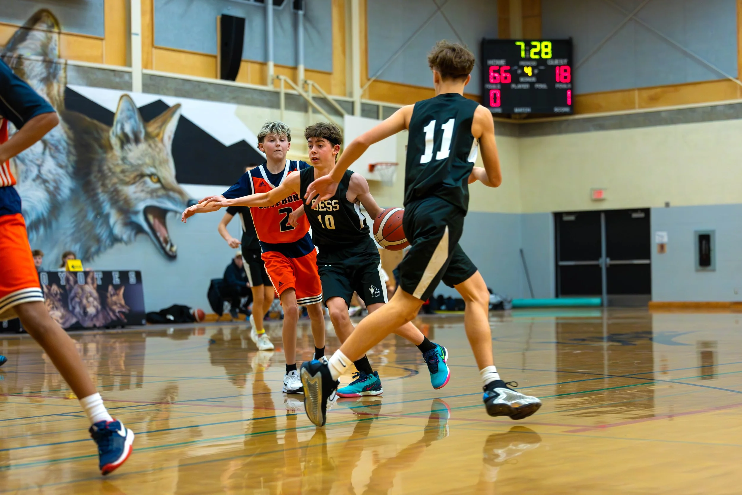 Young boys playing basketball in an indoor gym, with a wolf mural on the wall and a digital scoreboard showing 7:28 remaining.