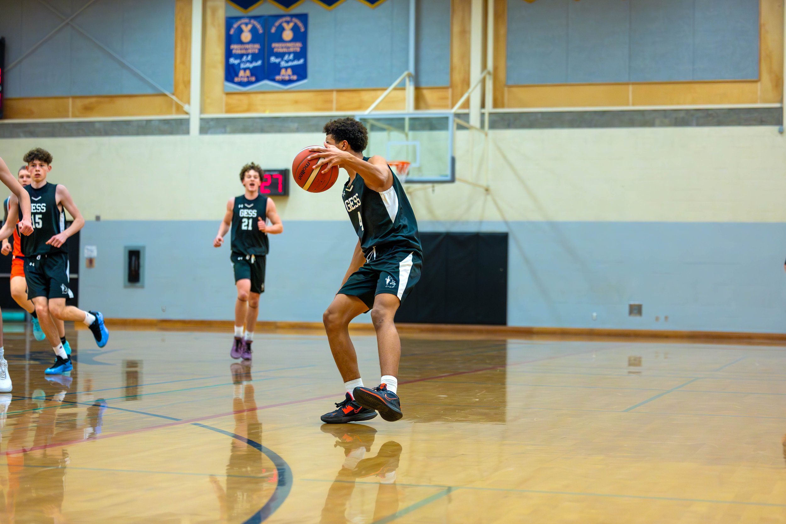 Young basketball player in black uniform holding a basketball in a gymnasium with teammates nearby.