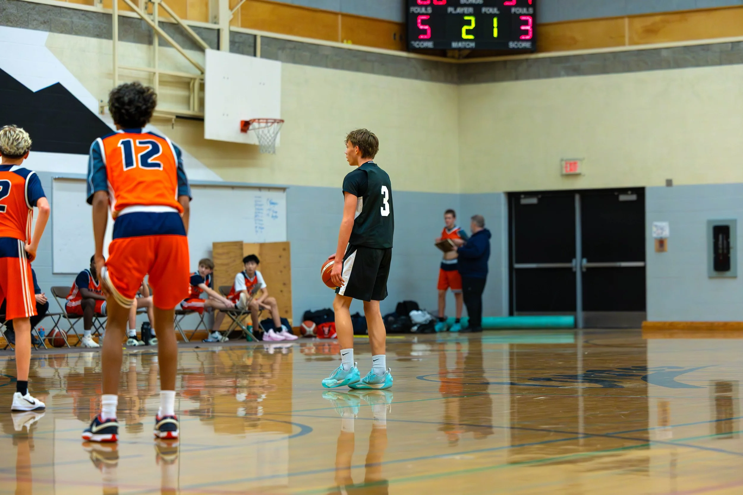 Young basketball player in black uniform holding a basketball on a gym court with orange team players; scoreboard showing 5-2-1-3; coaches and teammates sit on benches in the background.