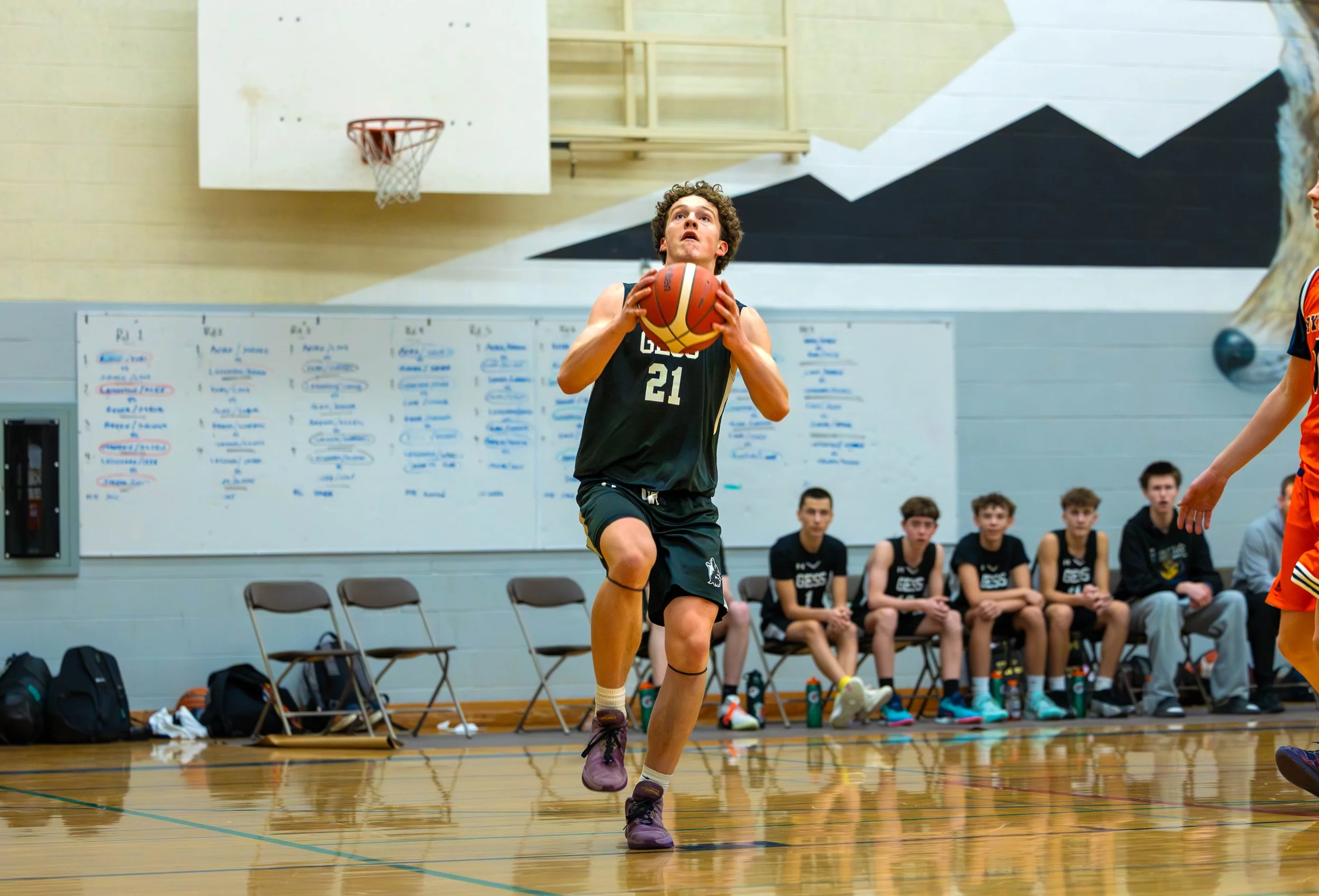 A young male basketball player in black jersey with number 21, holding a basketball, preparing to shoot inside a gymnasium with a whiteboard and seated teammates in the background.