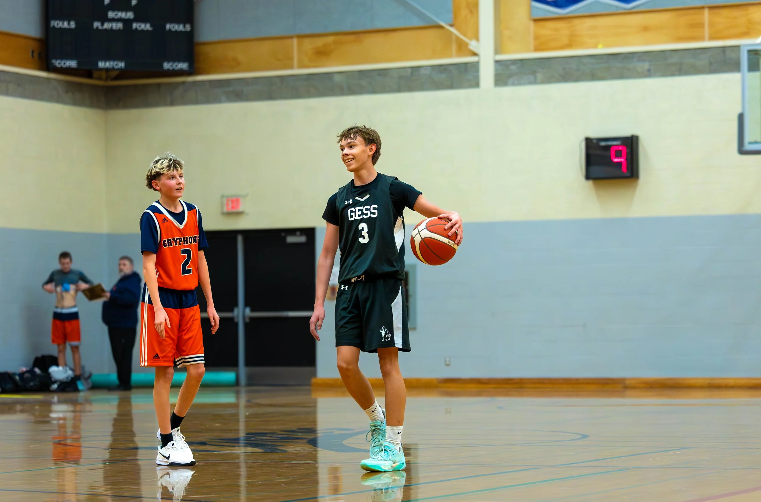 Two young male basketball players on an indoor court, one smiling with a basketball, and another looking at him, with scoreboard in the background.