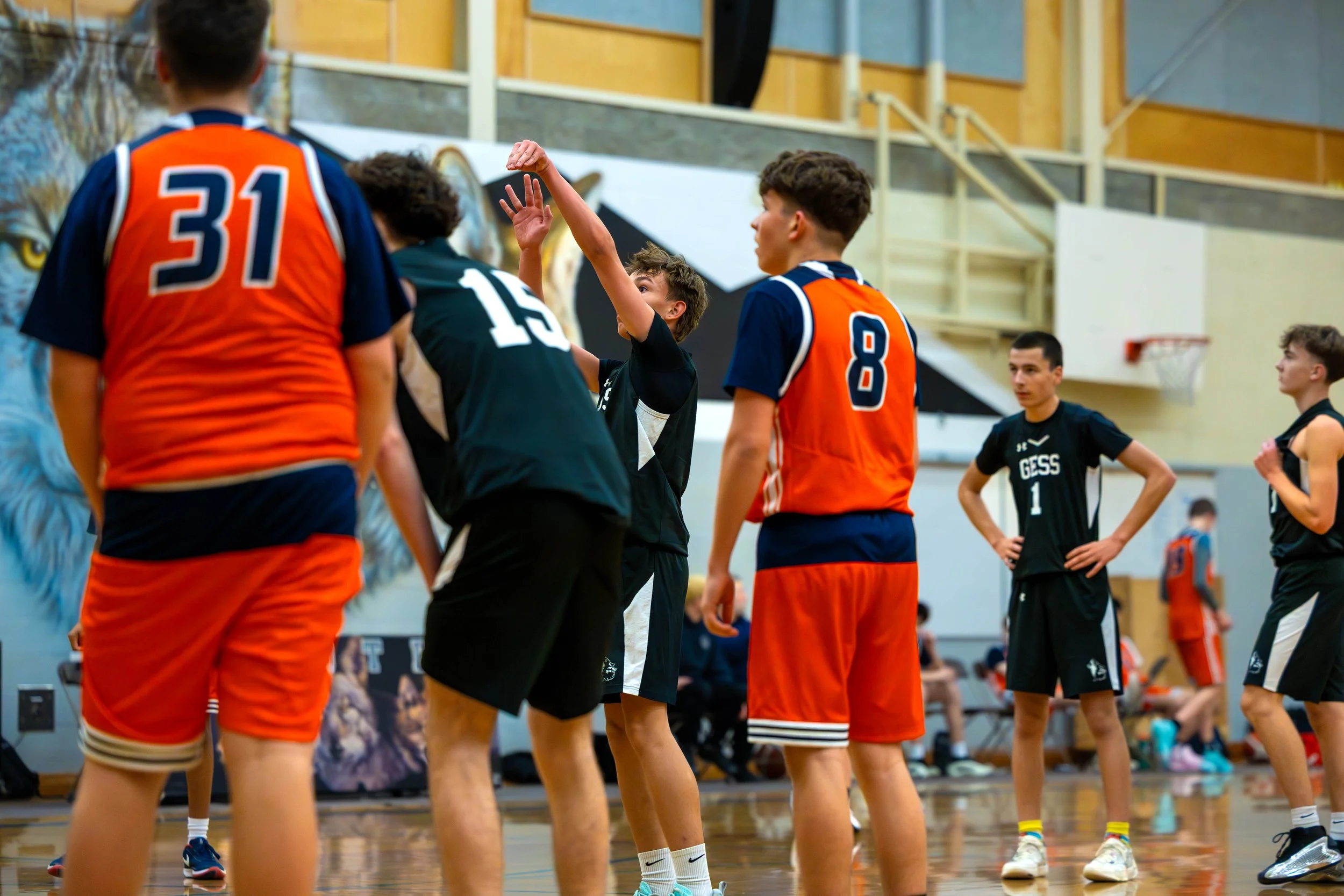 Young basketball players and a referee on an indoor court, some players wearing black jerseys and others in orange jerseys, with a mural of a lion in the background.