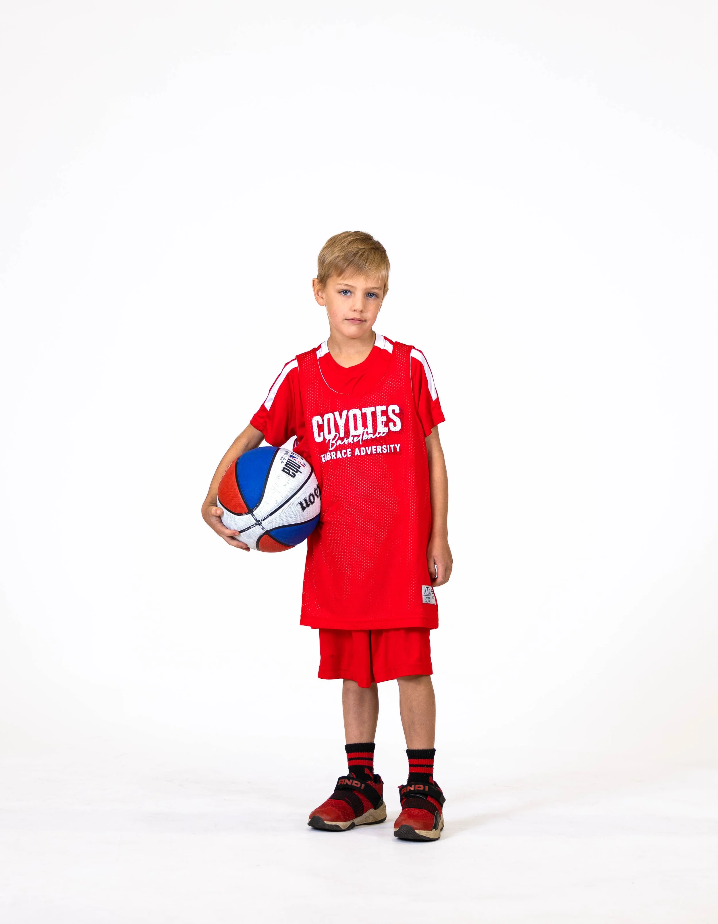 Young boy dressed in red basketball uniform holding a basketball, standing against a plain white background.
