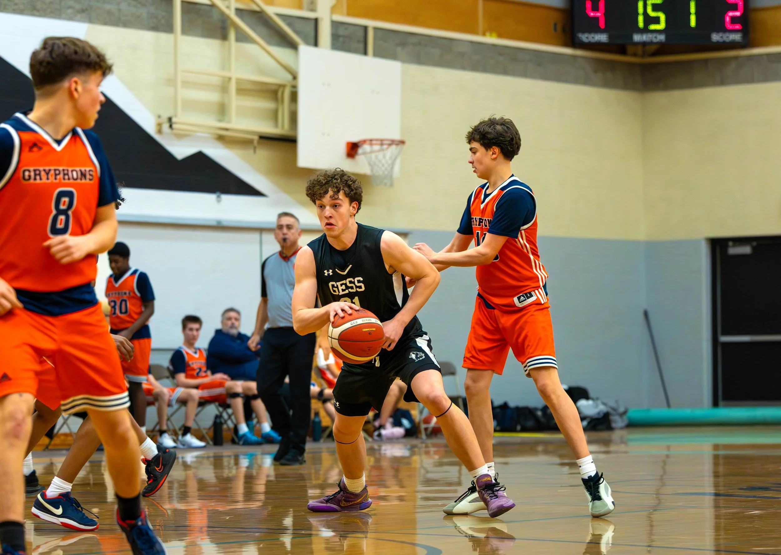 Young basketball players in action during a game in an indoor gym, with one player dribbling the ball while another attempts to block him. Teammates and referee are visible in the background.
