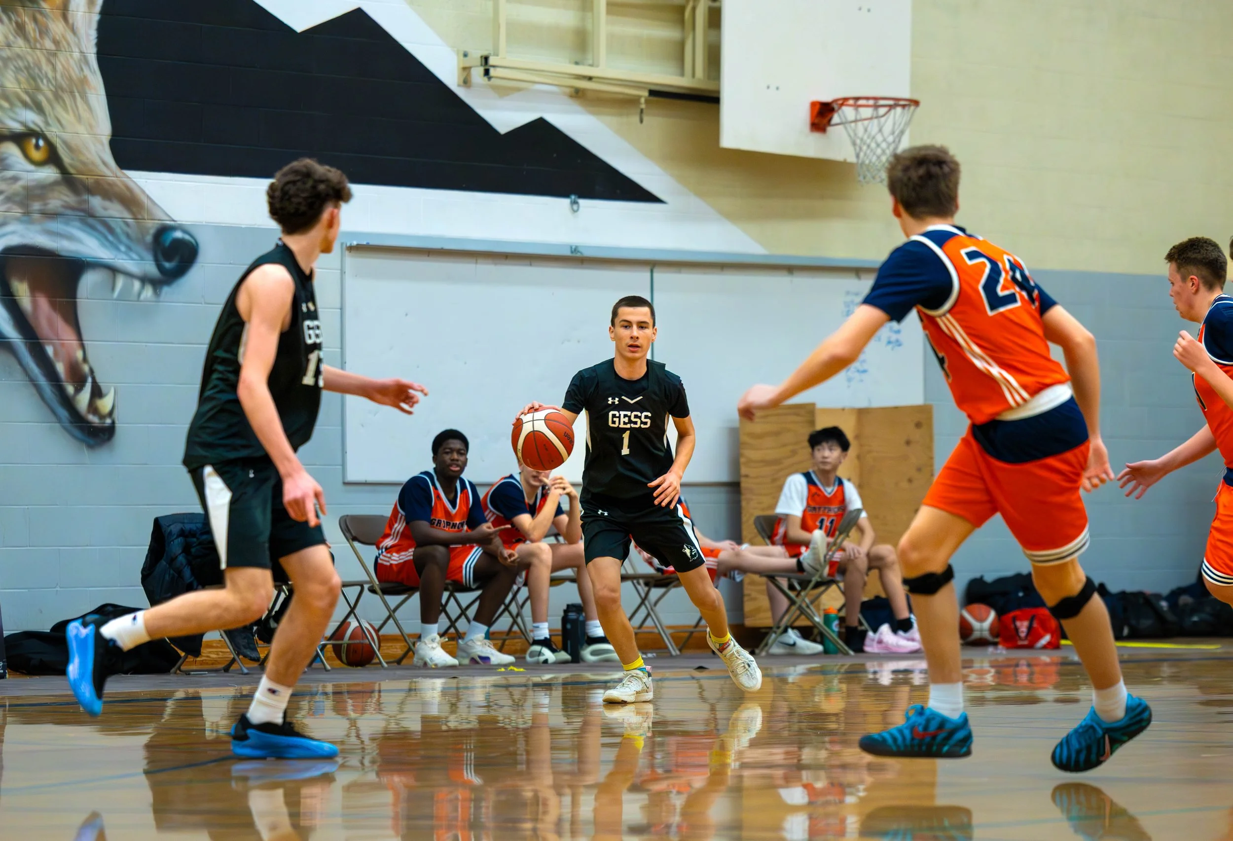 Young male basketball player in black team uniform dribbling the basketball on an indoor court, with teammates and opponents in the background, some sitting on chairs along the wall.