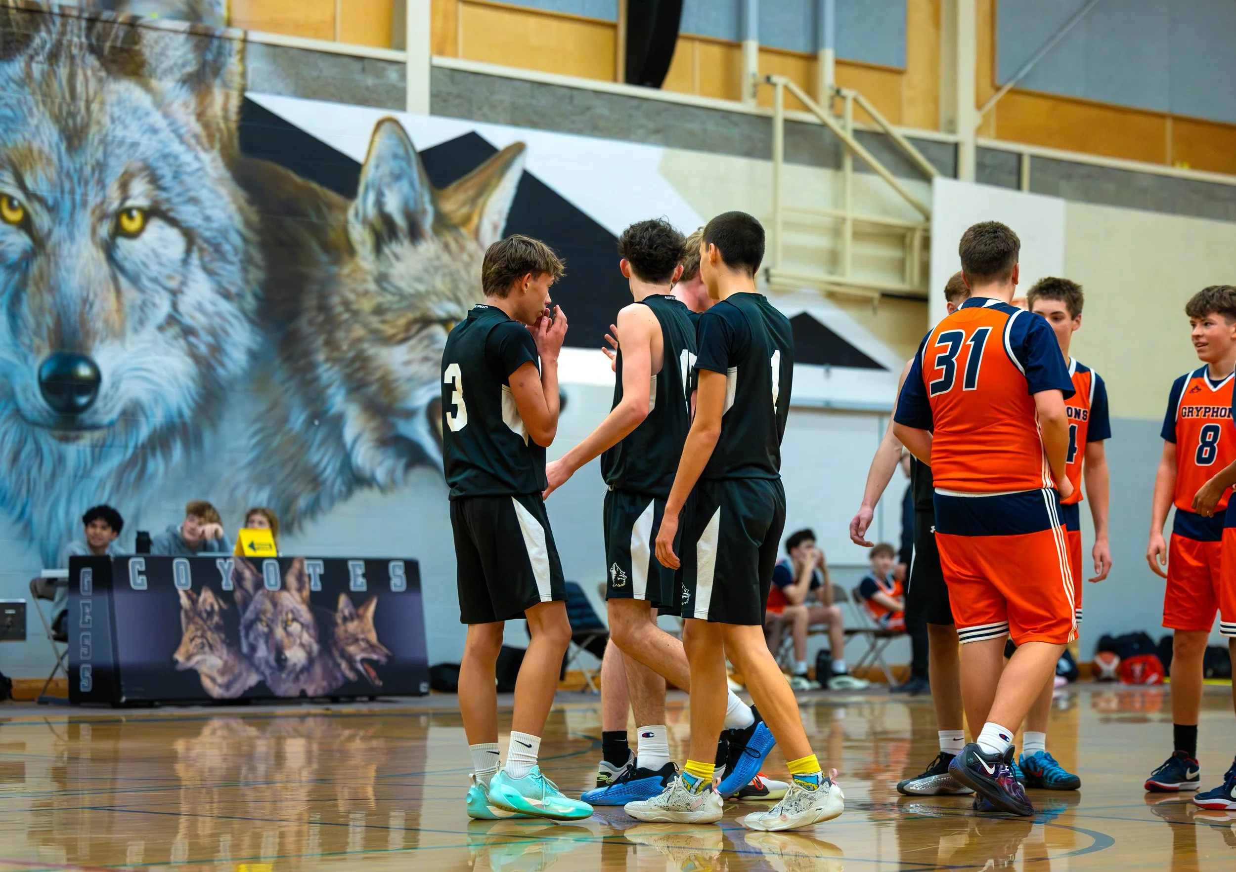 Youth basketball game in a gymnasium with a wolf mural in the background, players are gathered on the court, some standing and talking, others sitting on benches. Players are wearing black and orange jerseys.