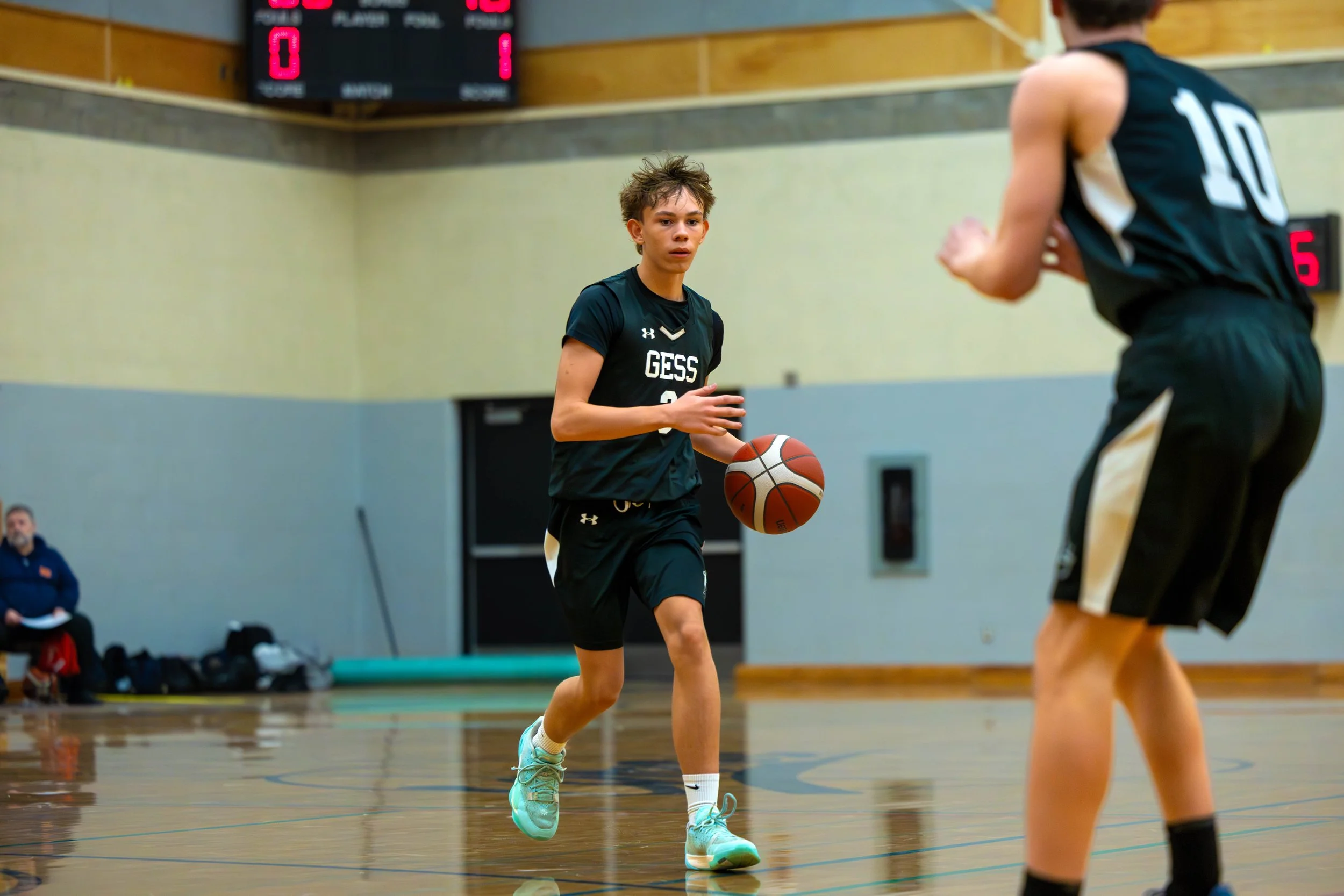 A young basketball player dribbling a basketball during a game in an indoor gym, with another player in a black uniform nearby and a digital scoreboard in the background.