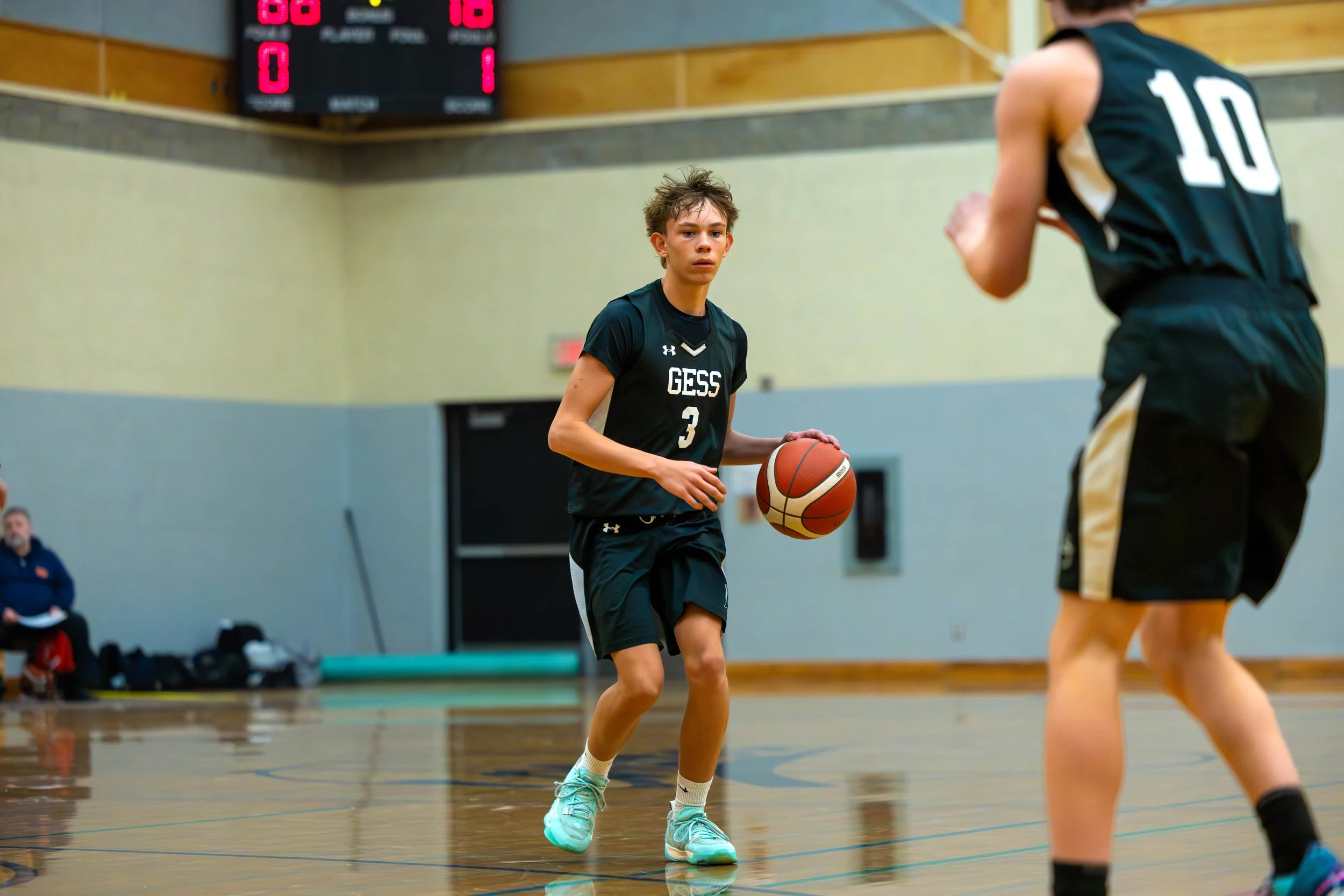 A young male basketball player in a black uniform with 'GESS' and number 3, dribbling a basketball on an indoor court, with another player and a scoreboard in the background.