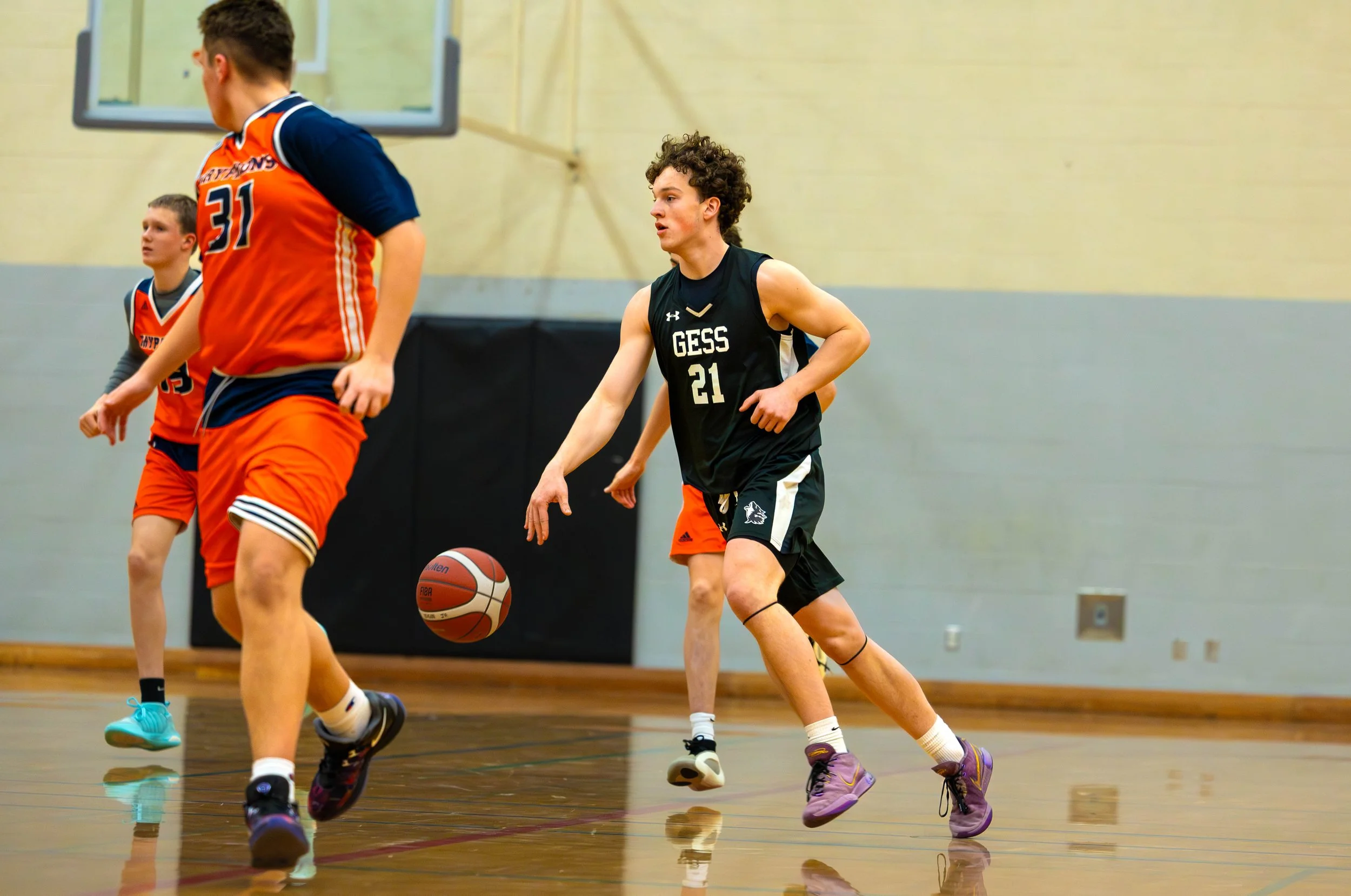 Young men playing basketball indoors, one wearing a black GESS jersey with the number 21, dribbling the ball while others in orange jerseys are defending.