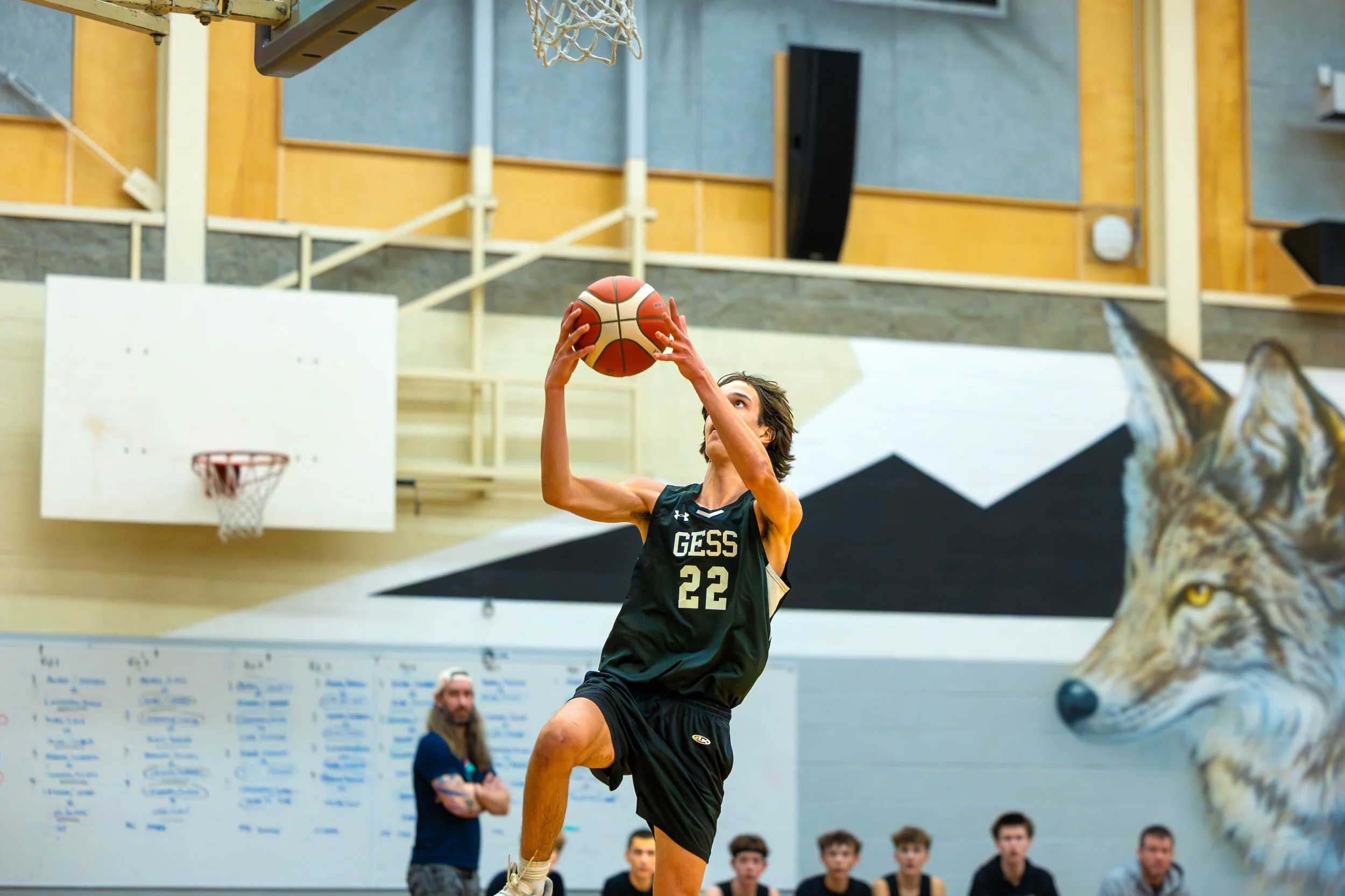 A basketball player in a black uniform with GESS 22 is jumping in the air to shoot a basketball inside a gymnasium, with a wolf mural on the wall and several people watching.