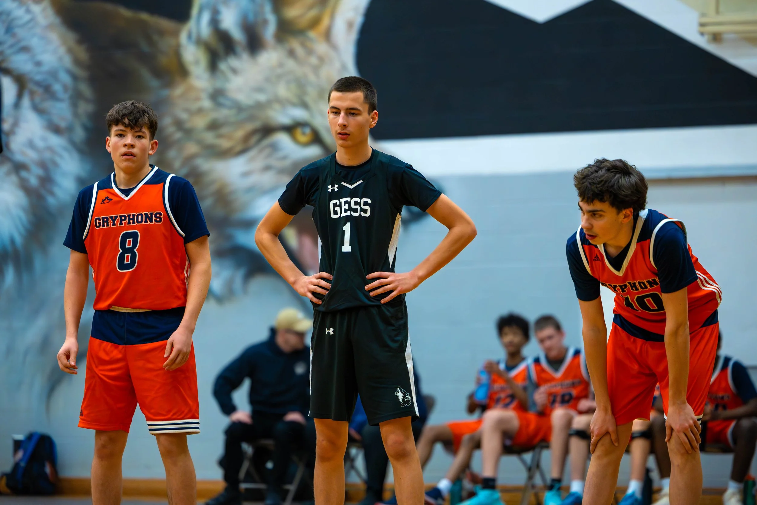 Three teenage boys wearing basketball uniforms standing on a gymnasium floor during a game. One is in an orange and blue uniform with the number 8, another in a black uniform with the number 1, and the third in orange and blue with the number 10. A l