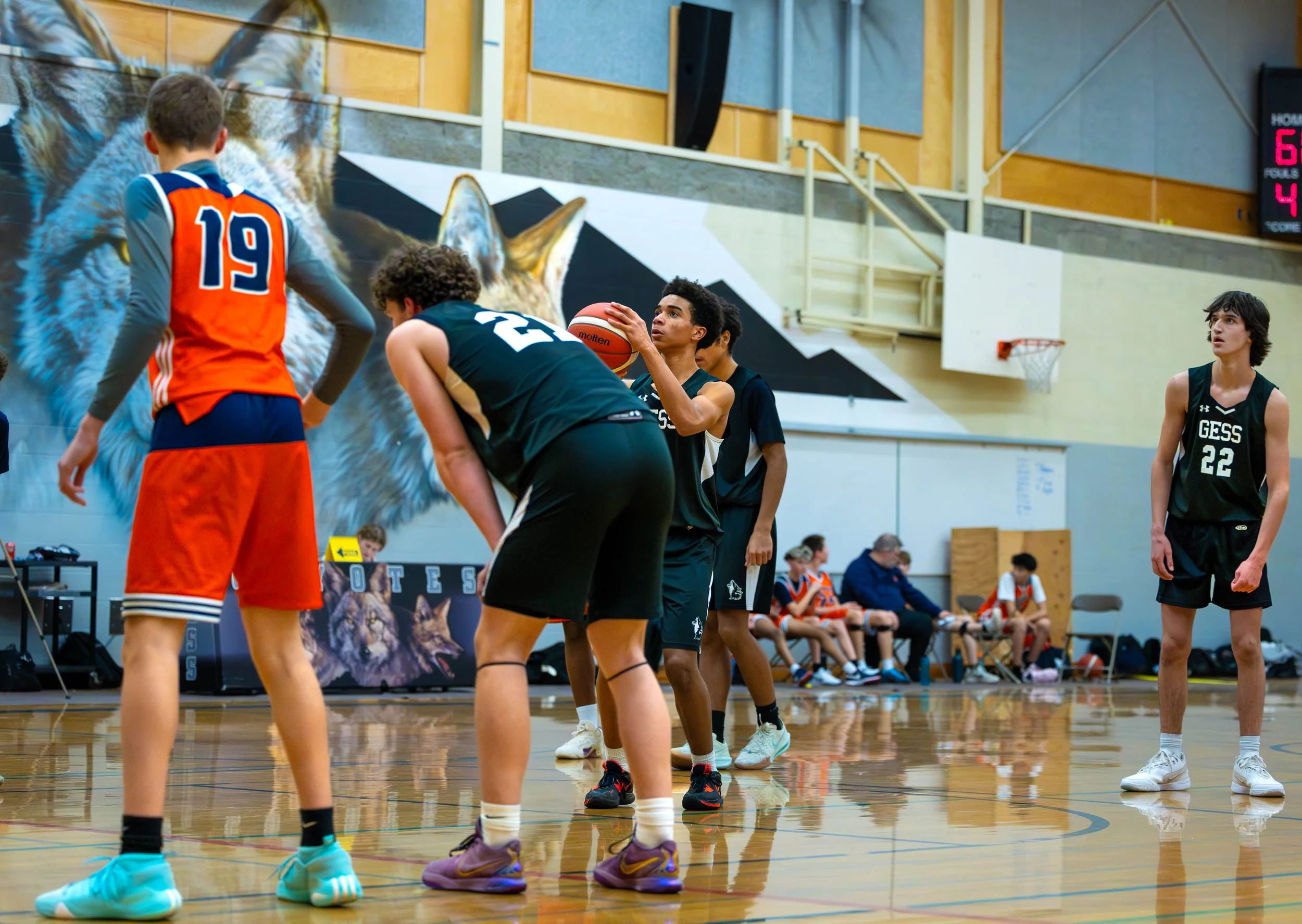 Young basketball players practicing free throws on an indoor court, with a mural of a wolf and a scoreboard in the background.