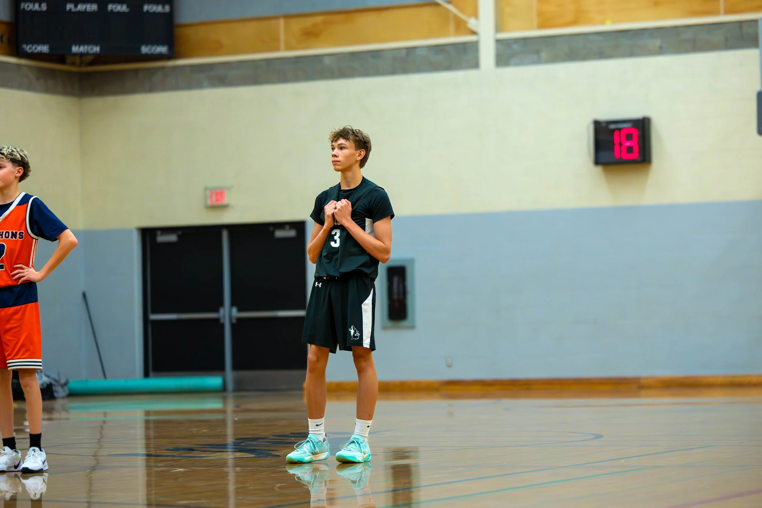 A young male basketball player standing on a gymnasium basketball court, wearing a black uniform with the number 3, holding his hands over his heart during a game.