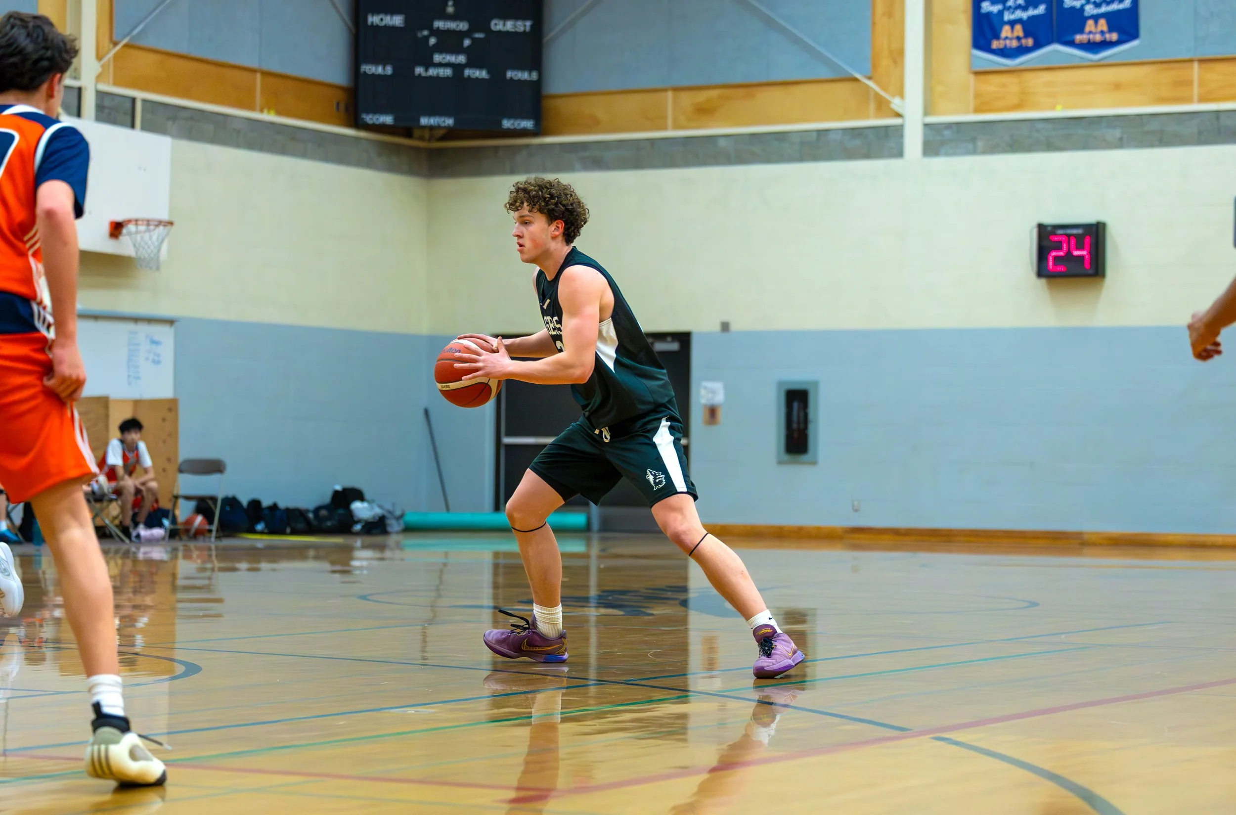 A young male basketball player with curly hair in a black uniform holding a basketball on a gymnasium court during a game.