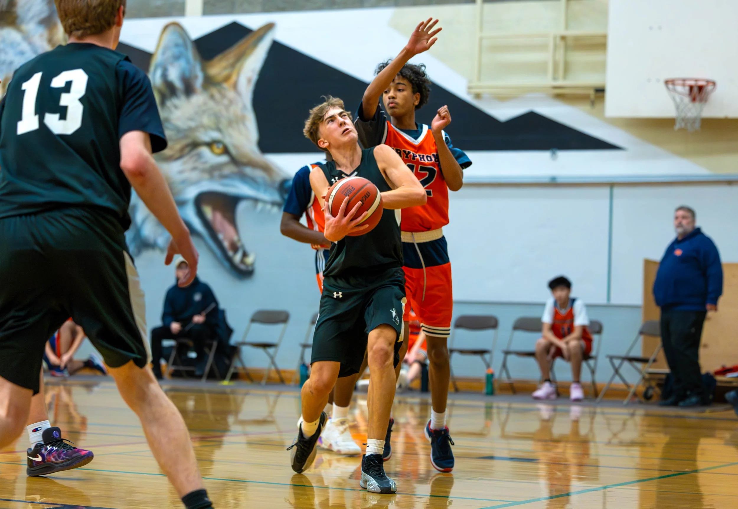 Young basketball player driving towards the hoop with a defender trying to block his shot during a game in an indoor gym.