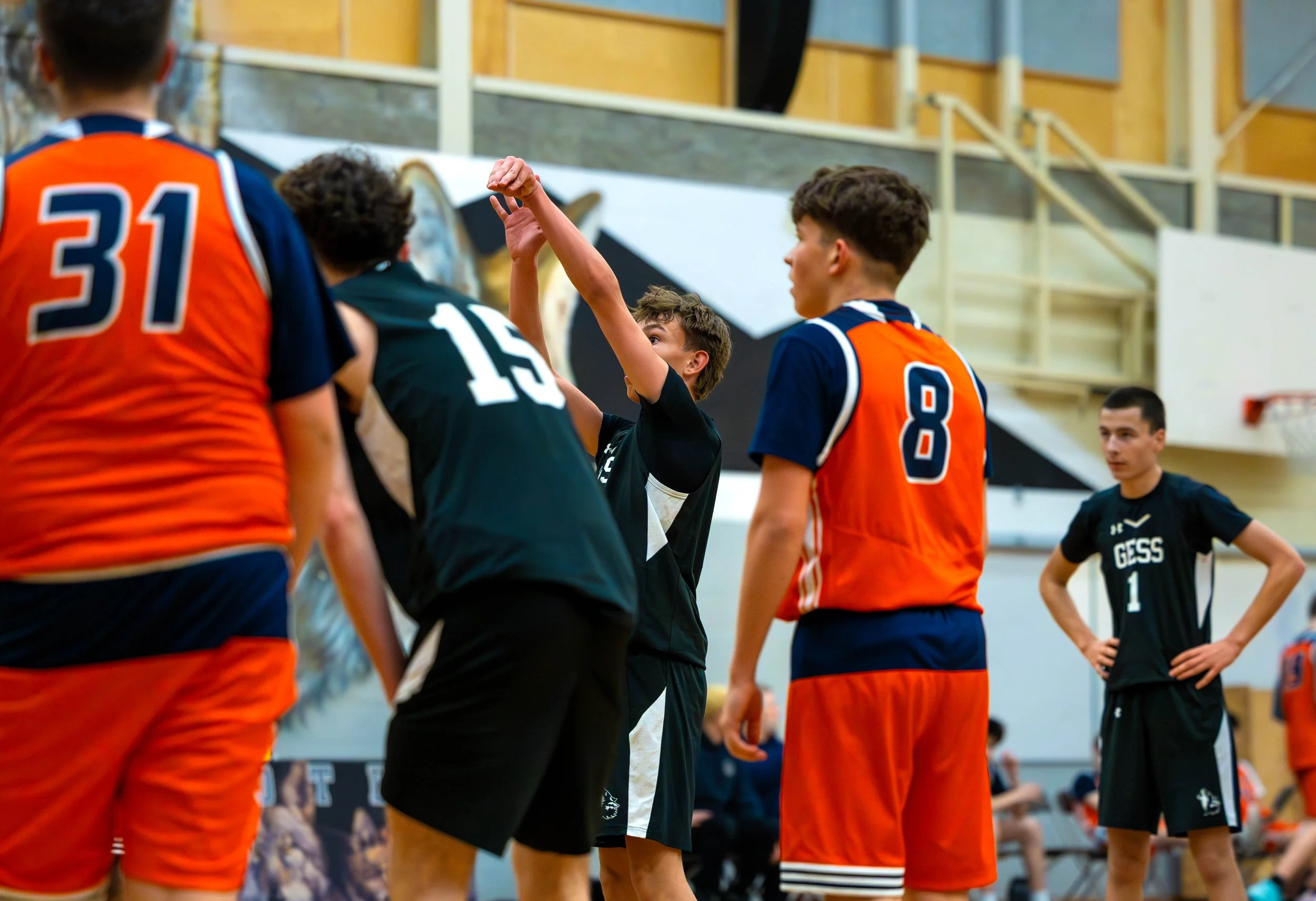 Young male basketball player in black jersey reaching up to make a shot while others watch in a gymnasium.