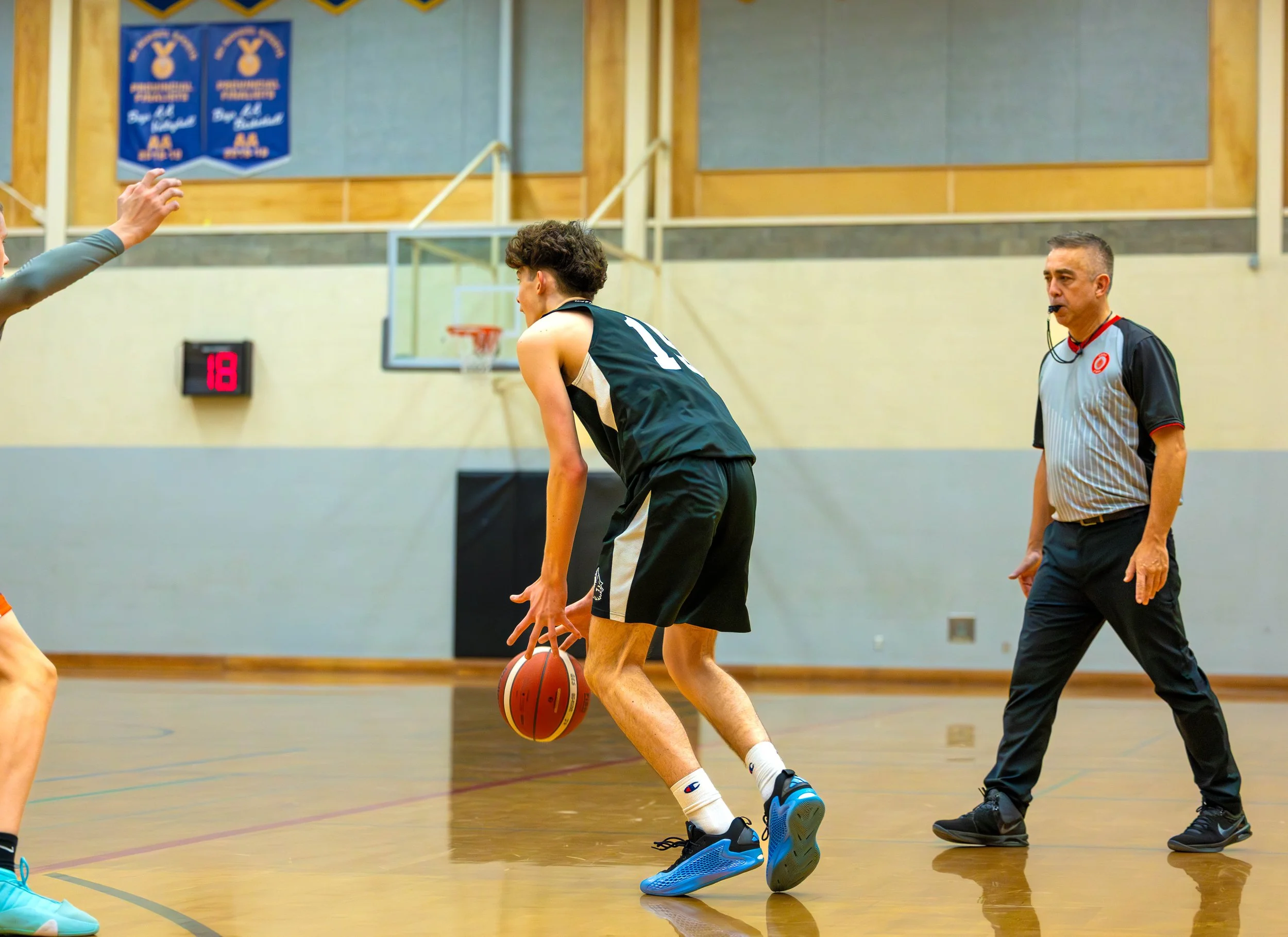 A basketball game in progress with a player dribbling the ball on the court, a referee standing nearby, and other players partially visible.