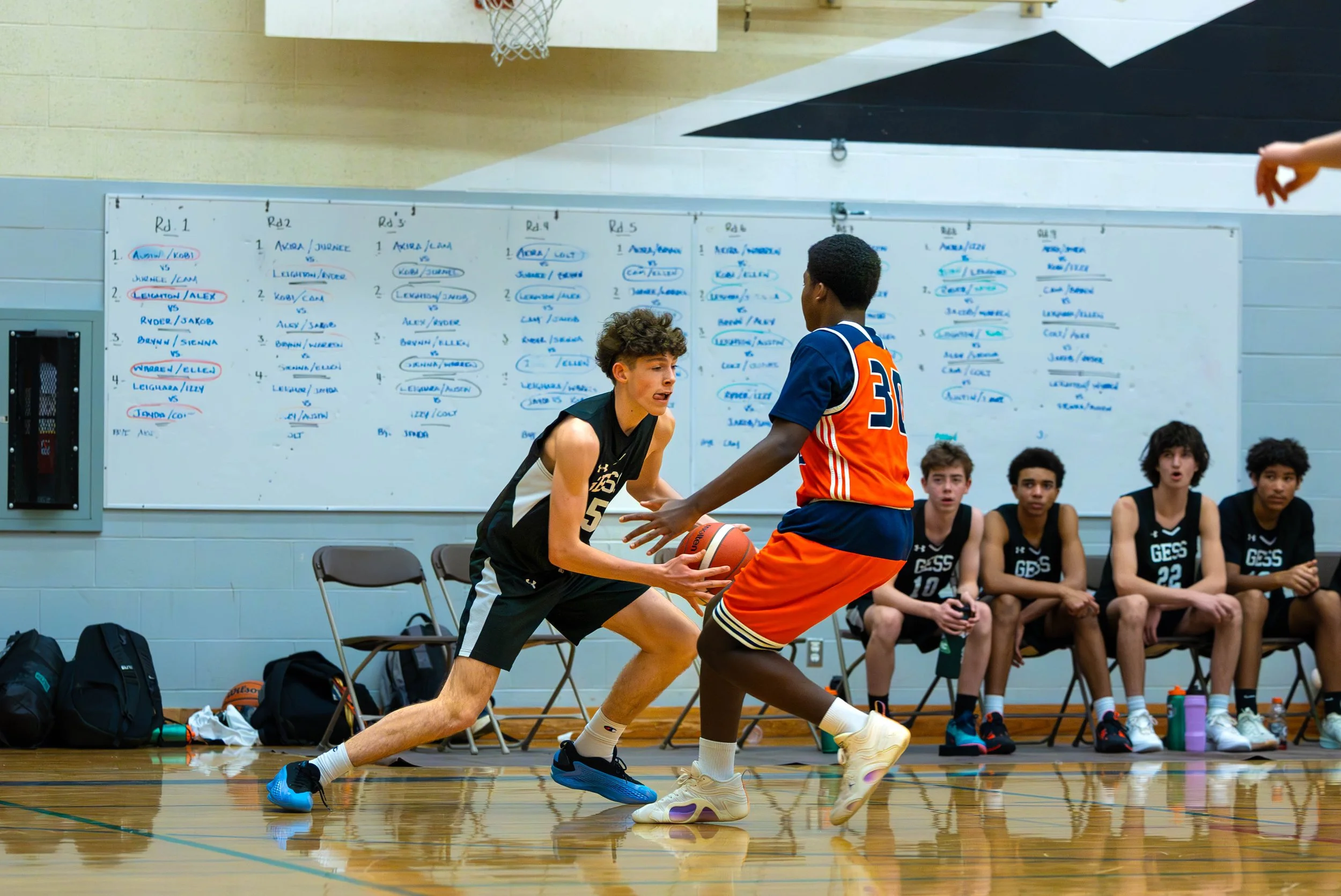 Two boys playing basketball on an indoor court with a whiteboard filled with notes and their teammates sitting on a bench in the background.