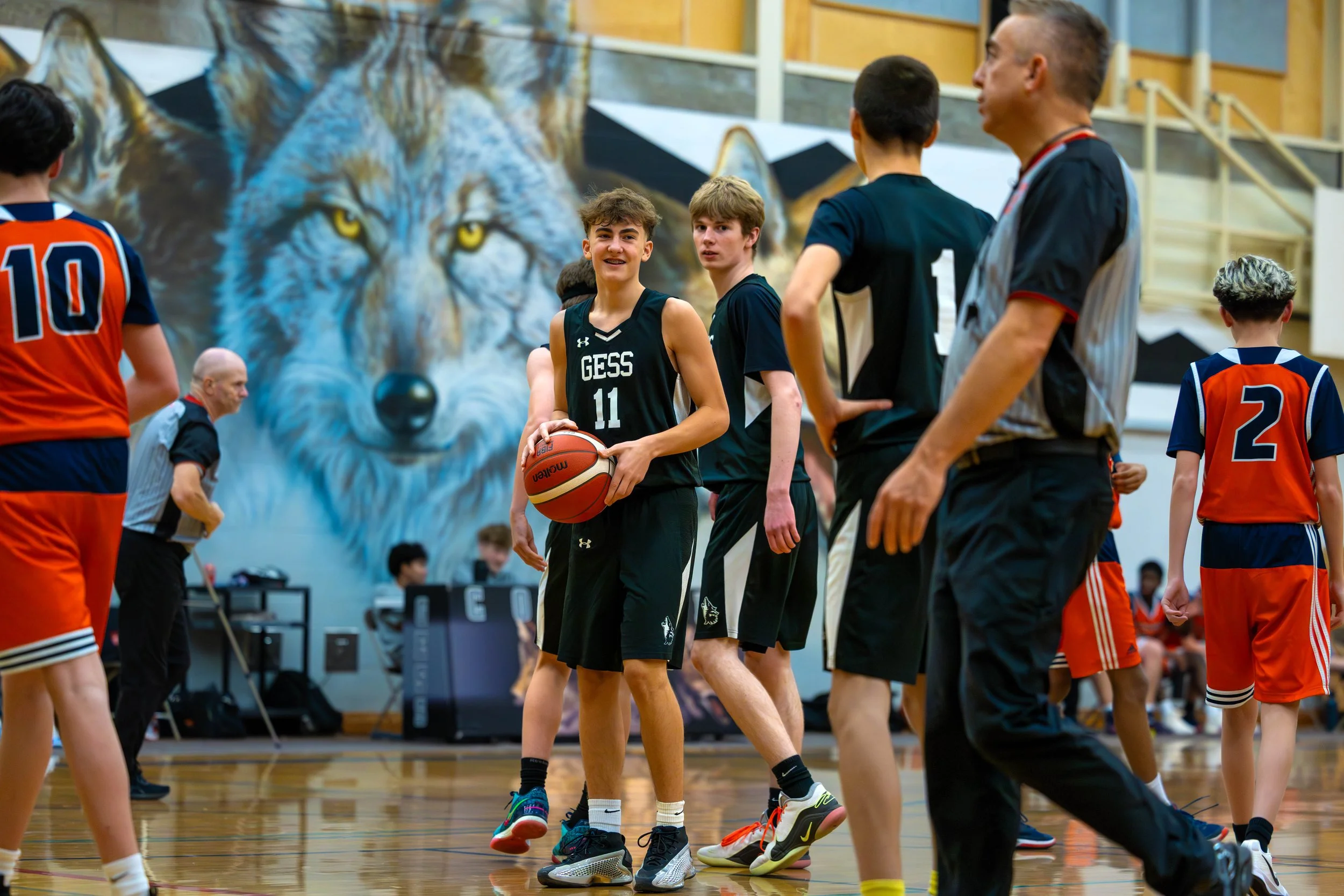 Youth basketball game in gym with players, referee, and a large wolf mural in background.