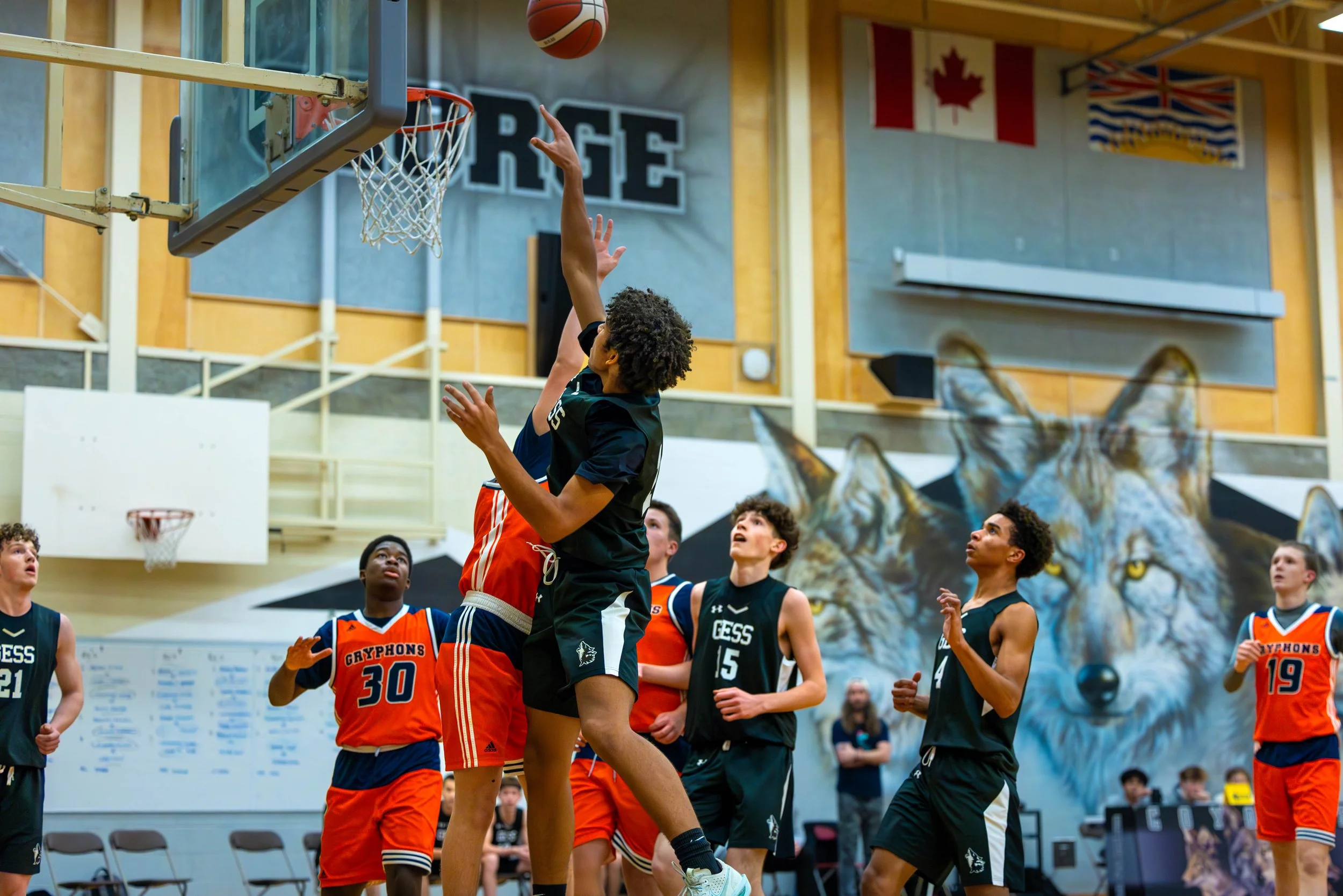 Young basketball players in black and orange jerseys playing in a gym, with a mural of a wolf in the background.