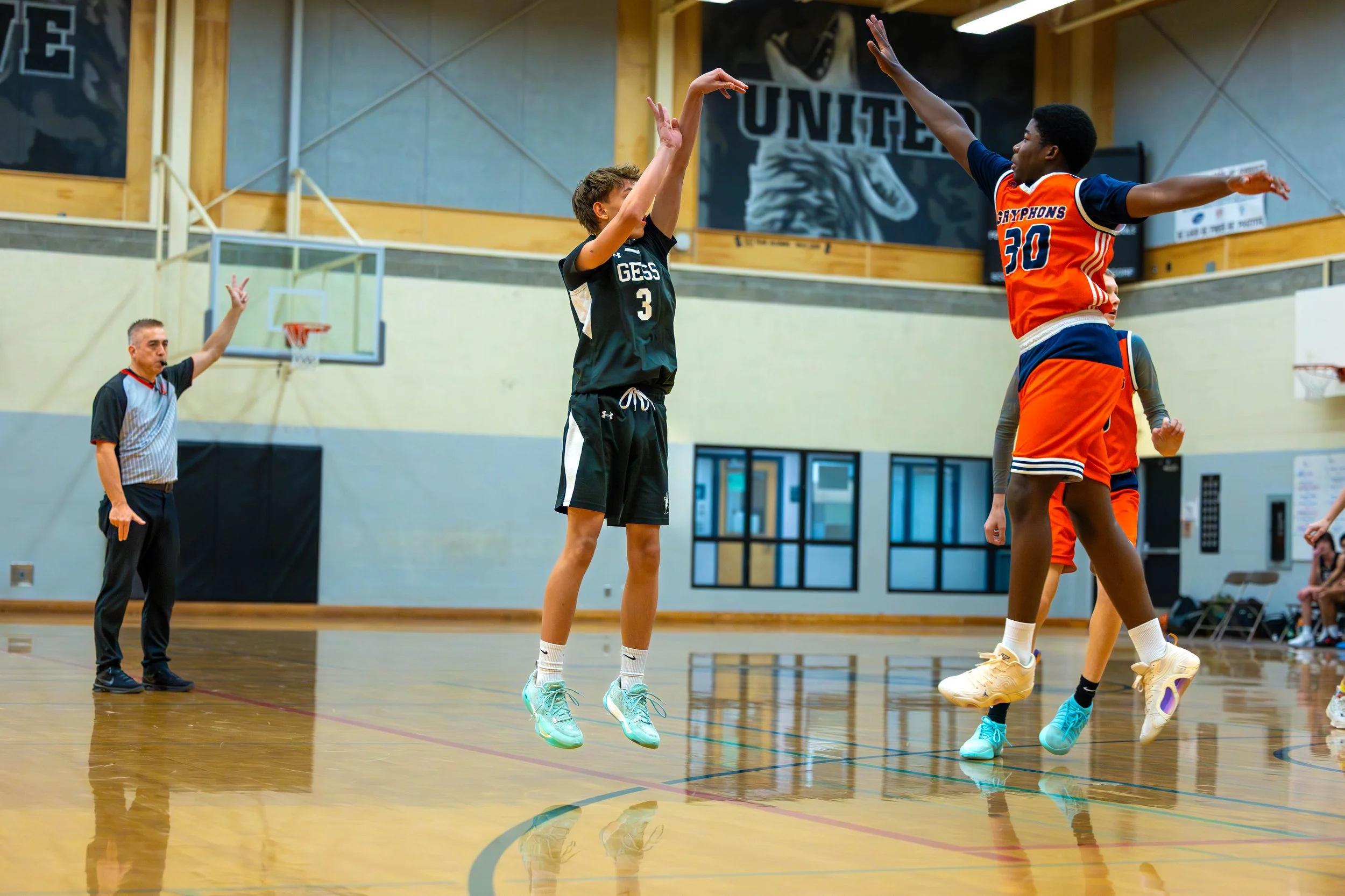 A basketball game shows two players jumping for the ball near the basket while a referee observes in the background, inside a gym with wood and gray walls, a large banner with a lion and 'UNITED' visible.