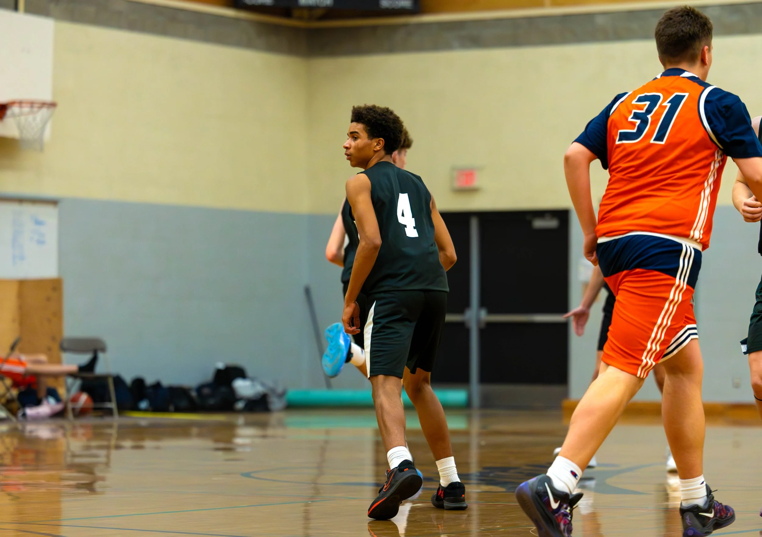 Young boys playing basketball indoors, one in a black uniform with the number 4, other in an orange and blue uniform with the number 31.