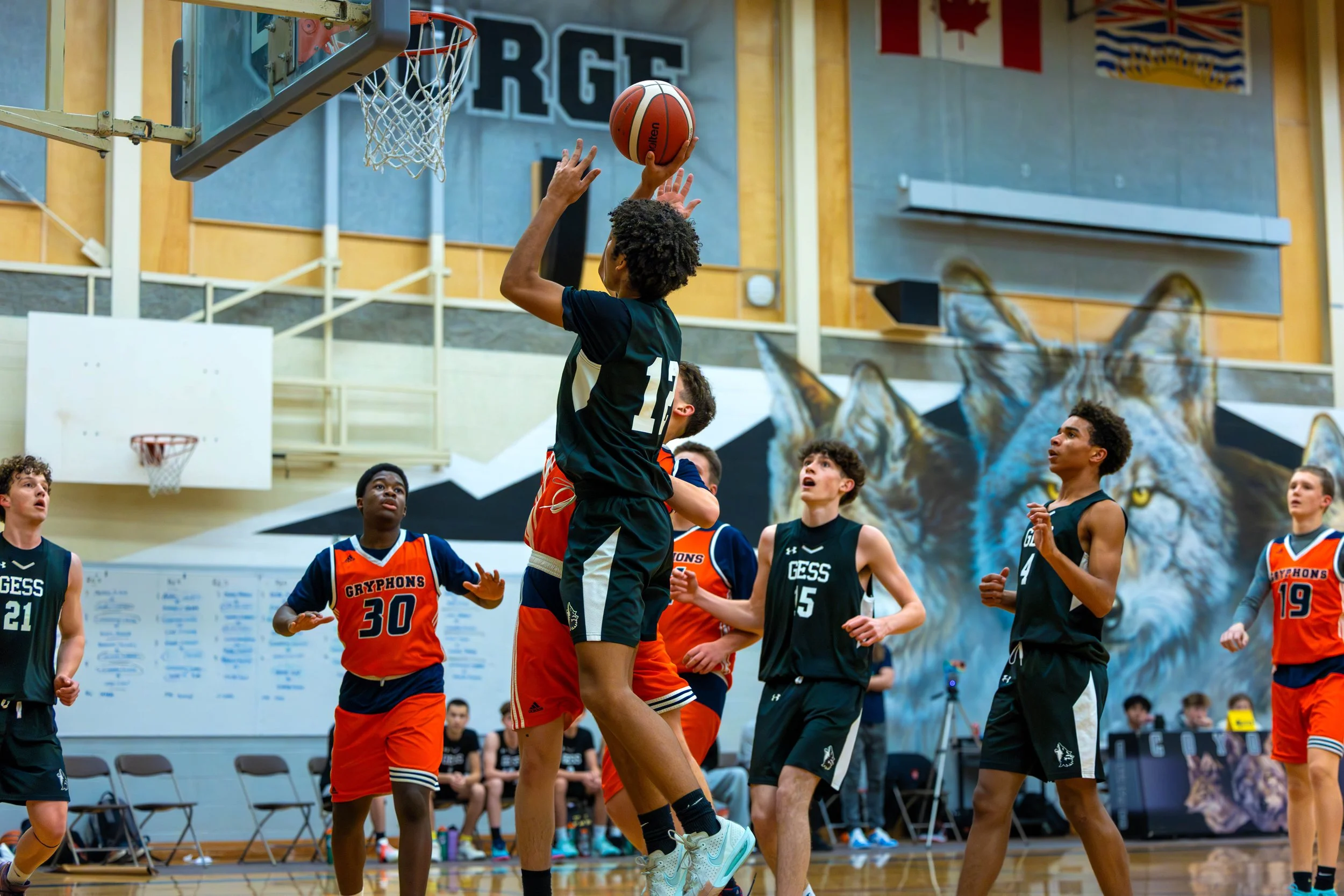 A basketball game with young players in black and orange jerseys inside a gym, with a mural of a wolf in the background.