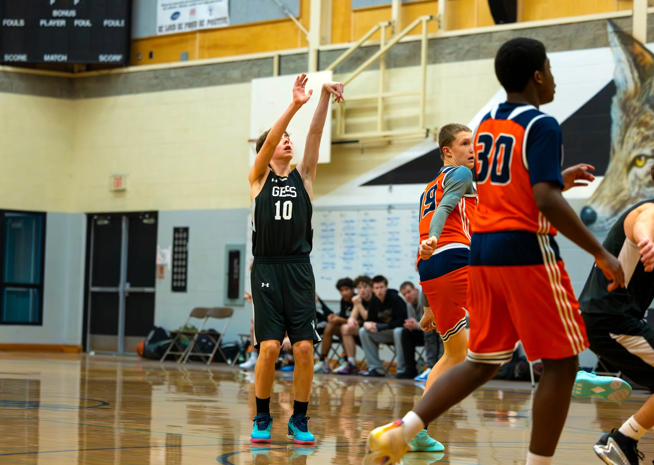 A youth basketball game in progress, with a player in black jersey shooting, and players in orange jerseys defending. The player in black is mid-shot, extending his arms, while the players in orange are moving on the court, with some looking towards 