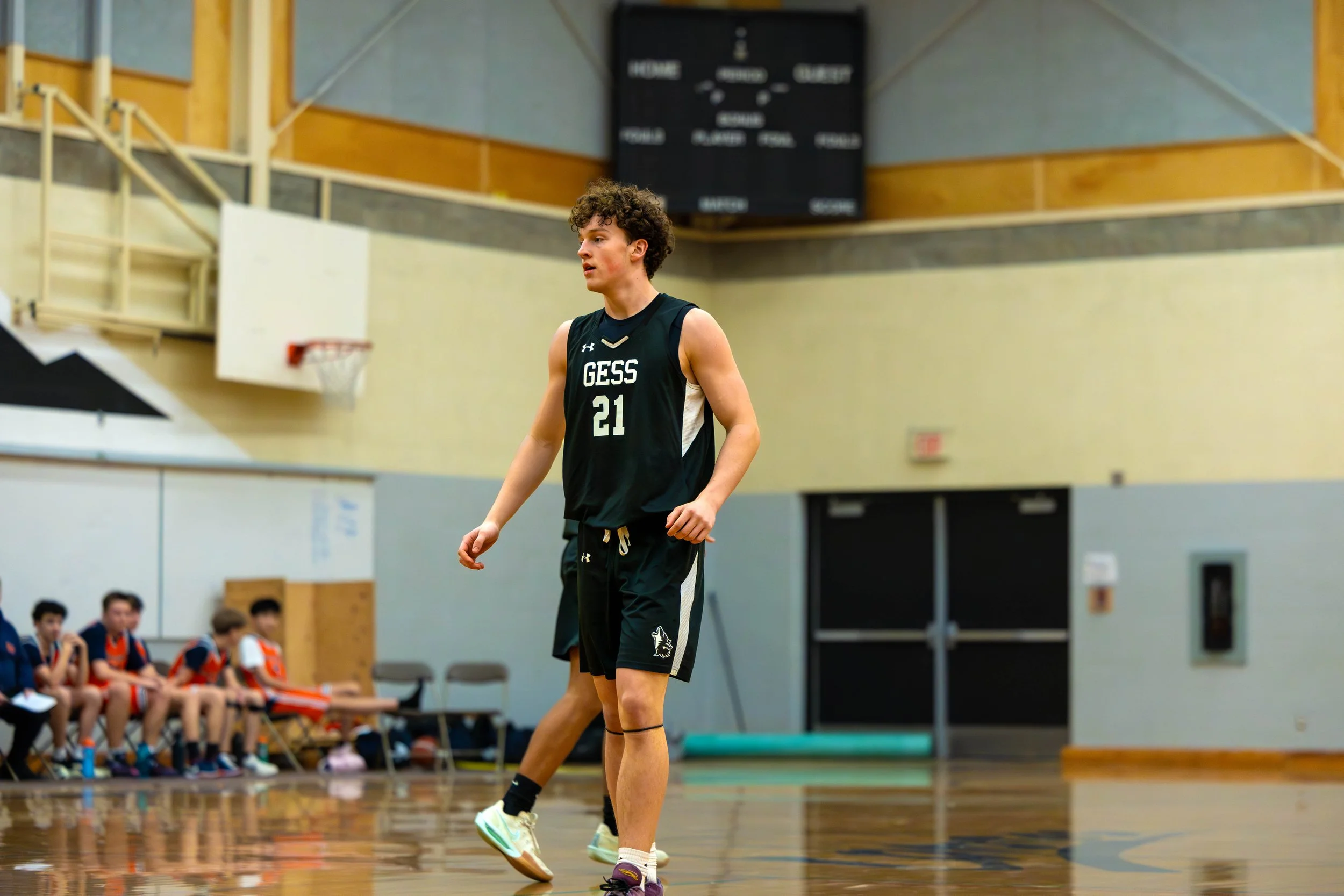 A young male basketball player in a black jersey with 'GESS' and the number 21, standing on an indoor basketball court, with a game scoreboard and teammates sitting on the bench in the background.
