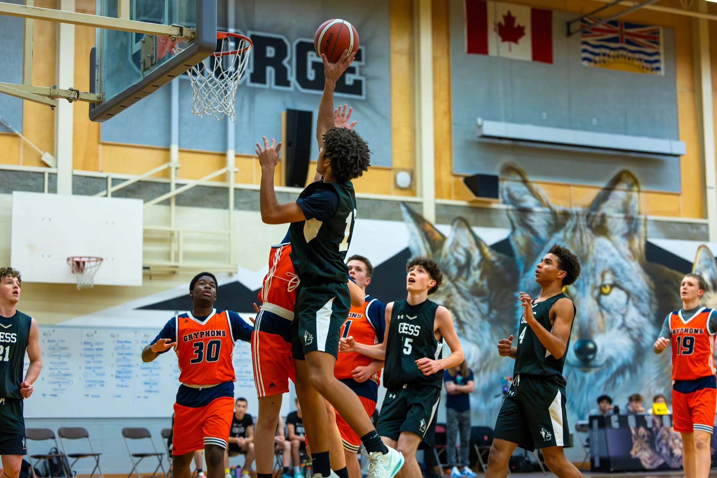Young basketball players in action during a game, with one player jumping to make a shot, in a gym with wolf mural on the wall and flags hanging from the ceiling.