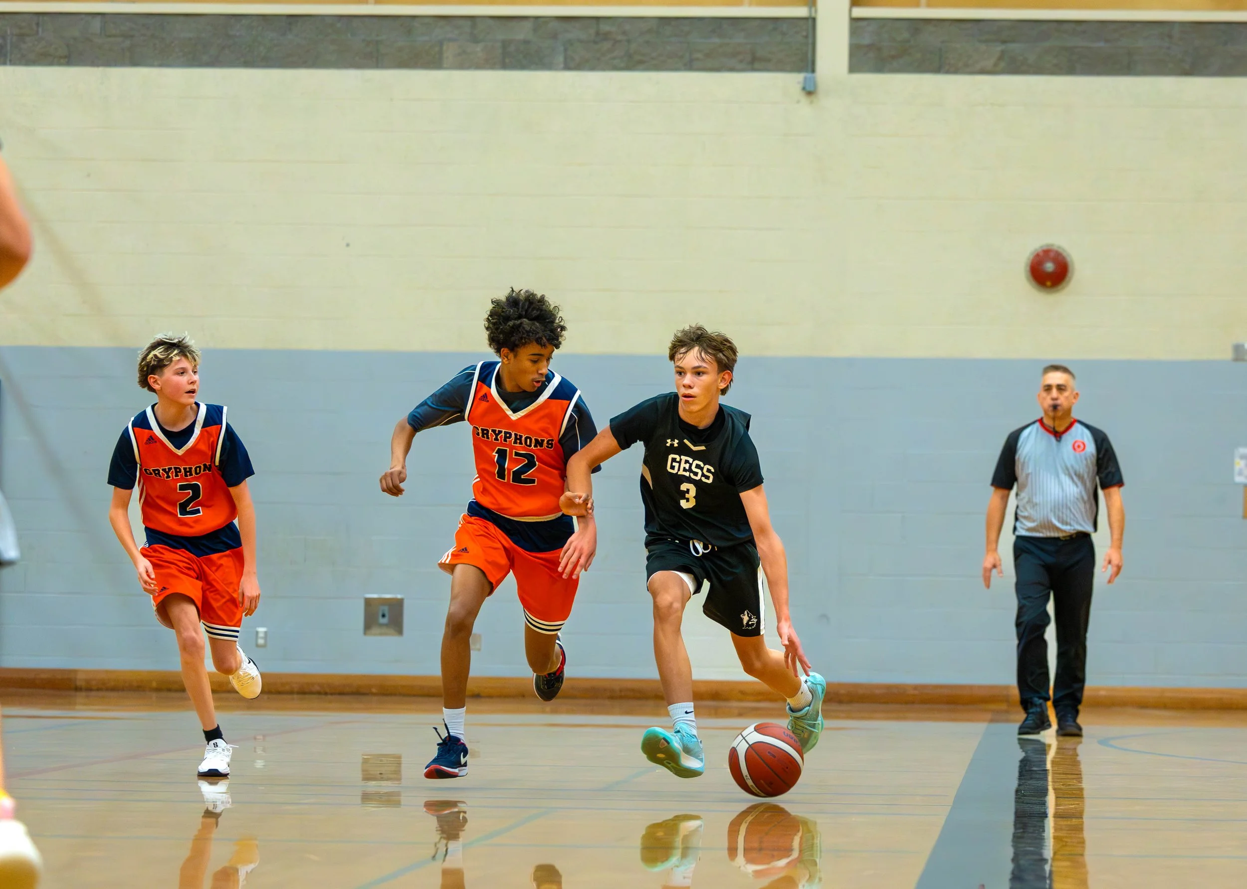 Kids playing basketball inside a gym, with an adult referee watching.
