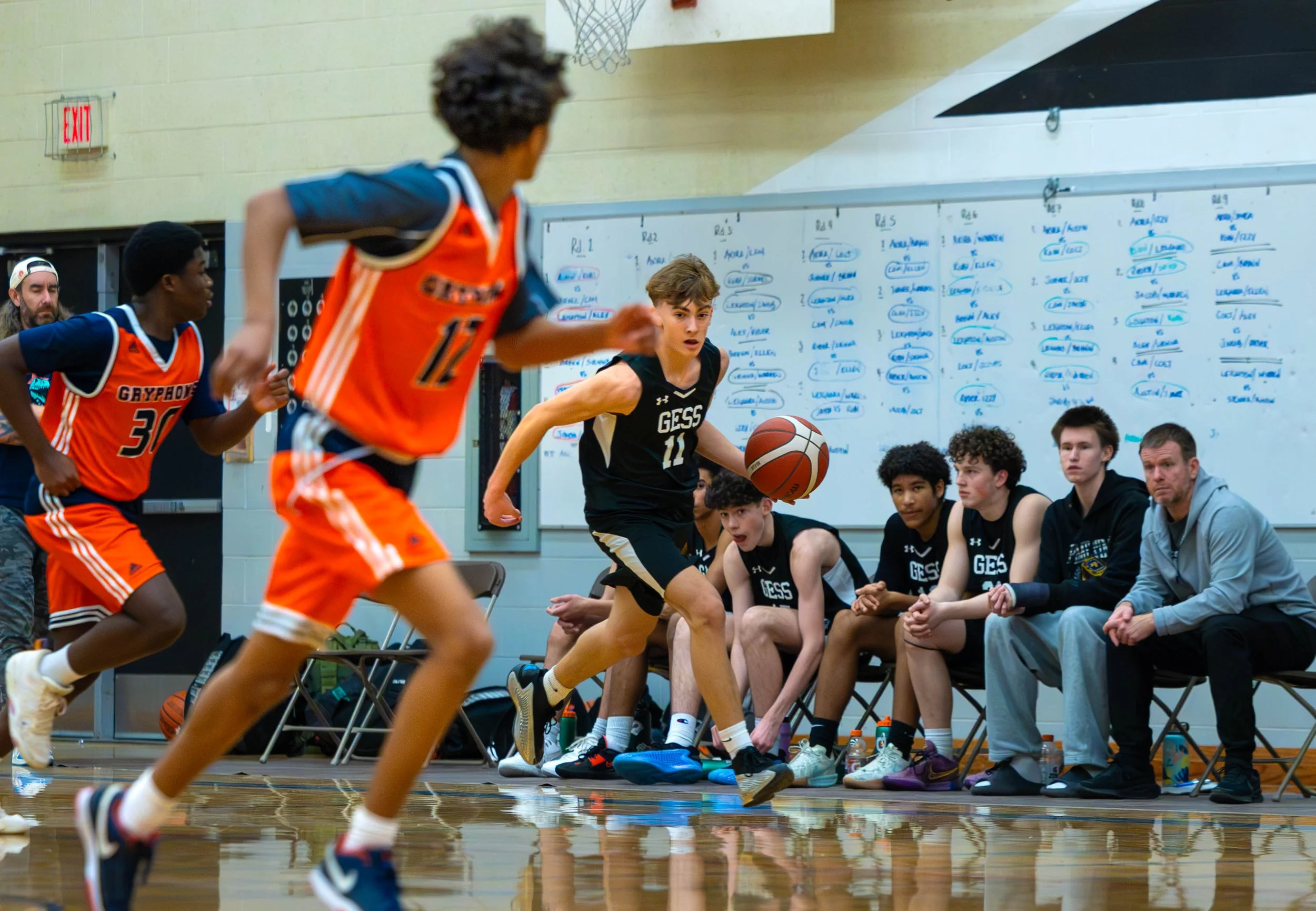 Young basketball players in action during a game, with players from one team wearing orange jerseys and the other team in black jerseys, while team members and coaches sit on the bench along the side of a gymnasium with a whiteboard in the background