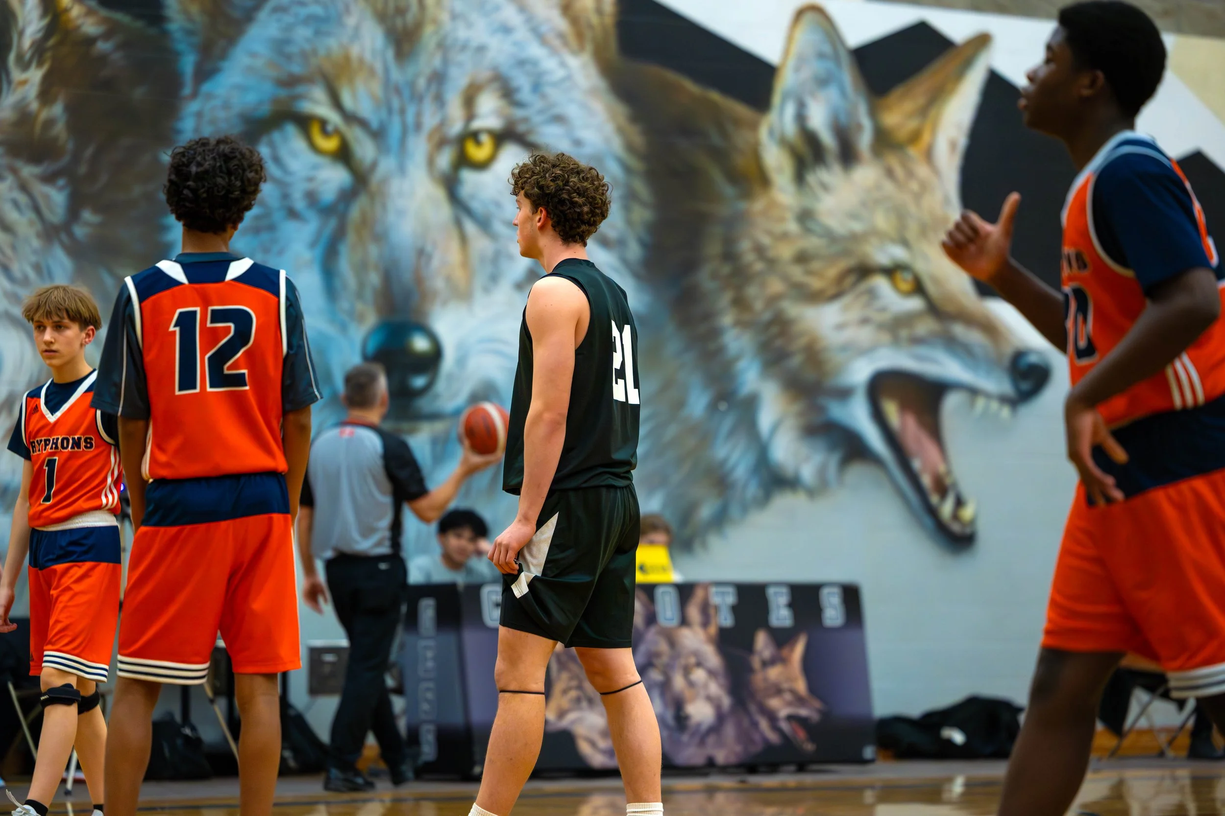Young basketball players in orange and black uniforms on the court during a game, with a large mural of wolves on the wall in the background.