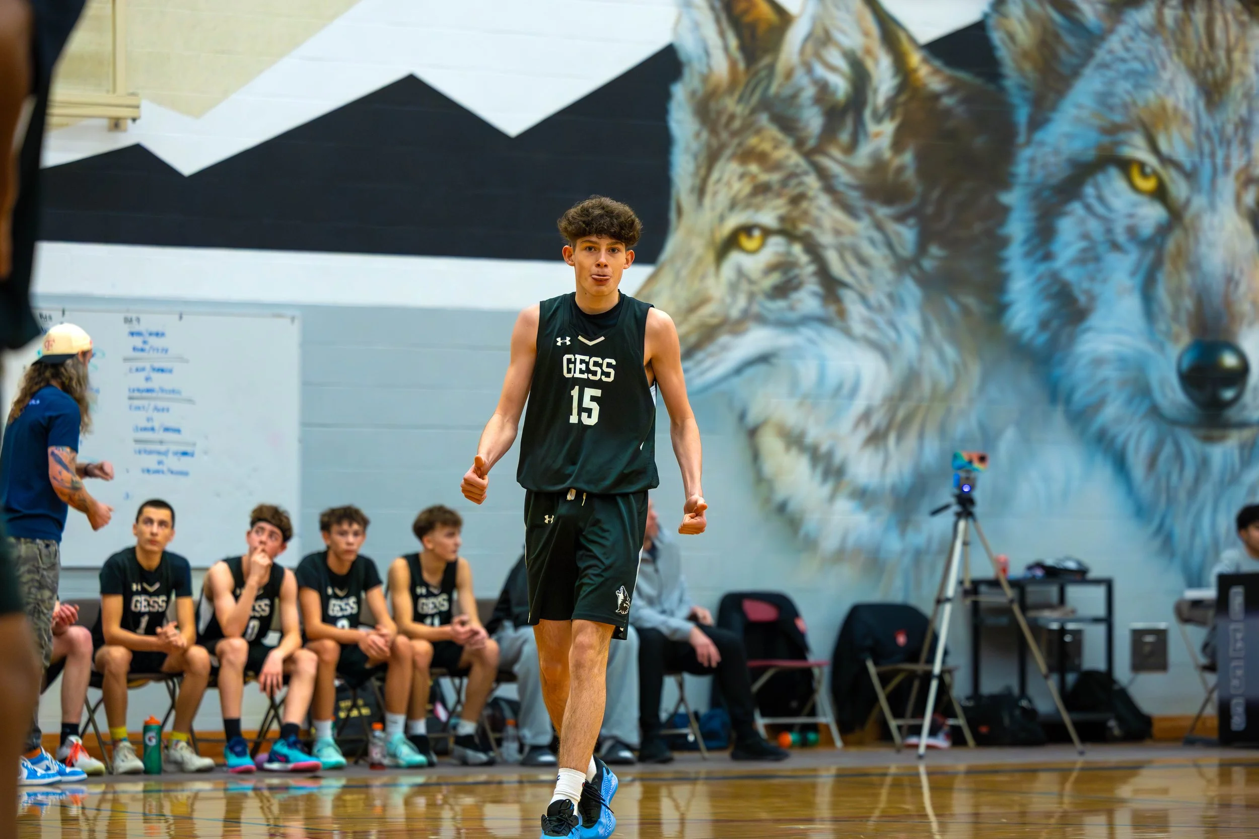 A youth basketball player wearing a black jersey with 'GESS' and the number 15, standing on a basketball court with a large wolf mural in the background, and teammates sitting on the bench behind.