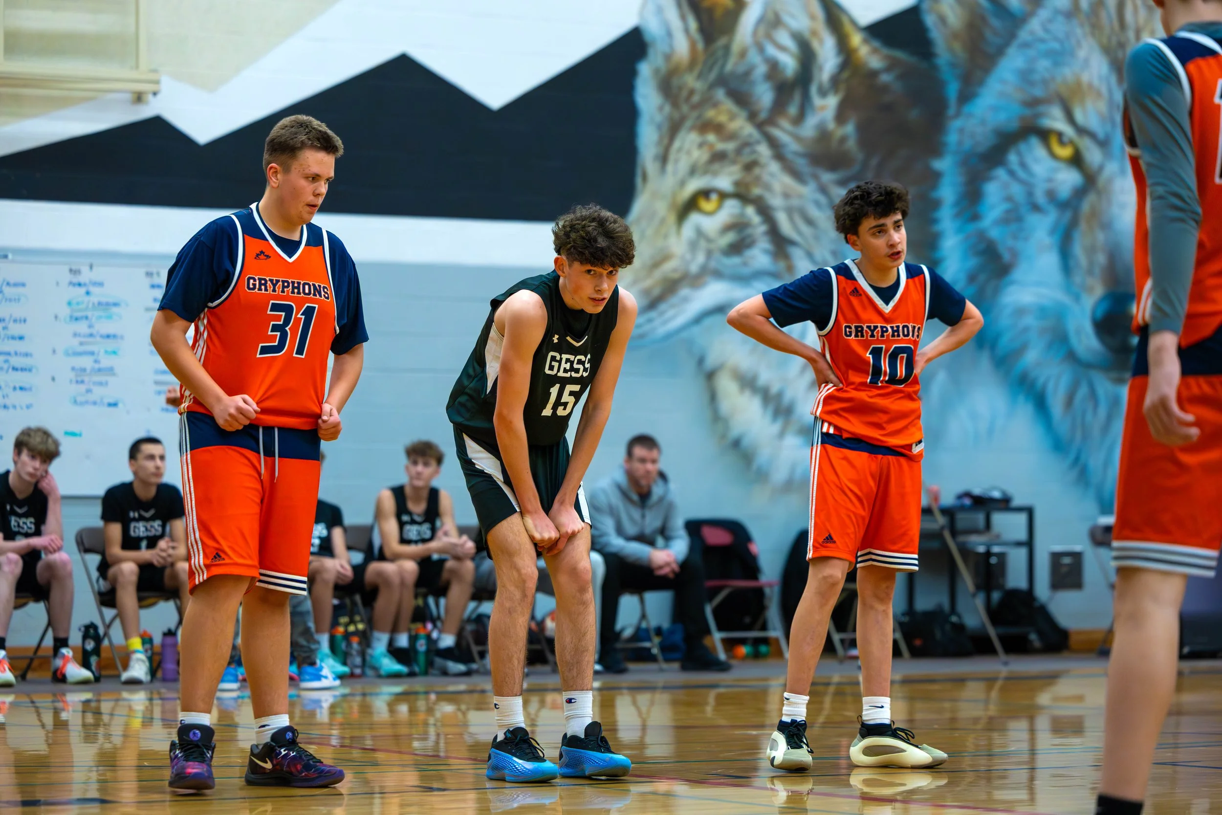 Young male basketball players in a gym, with some sitting on the bench and others standing, wearing jerseys with team names 'Gryphons' and 'GESS'. One player appears to be standing and looking down.