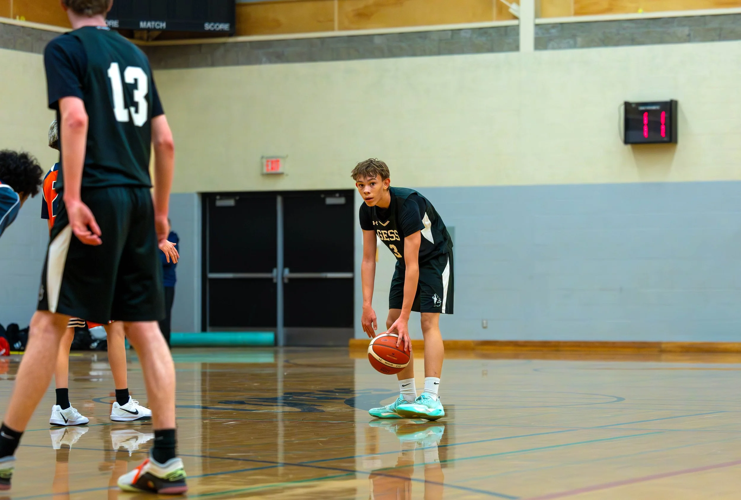 Young basketball player in black uniform standing on a basketball court, holding a basketball and preparing to take a shot, with other players around him.