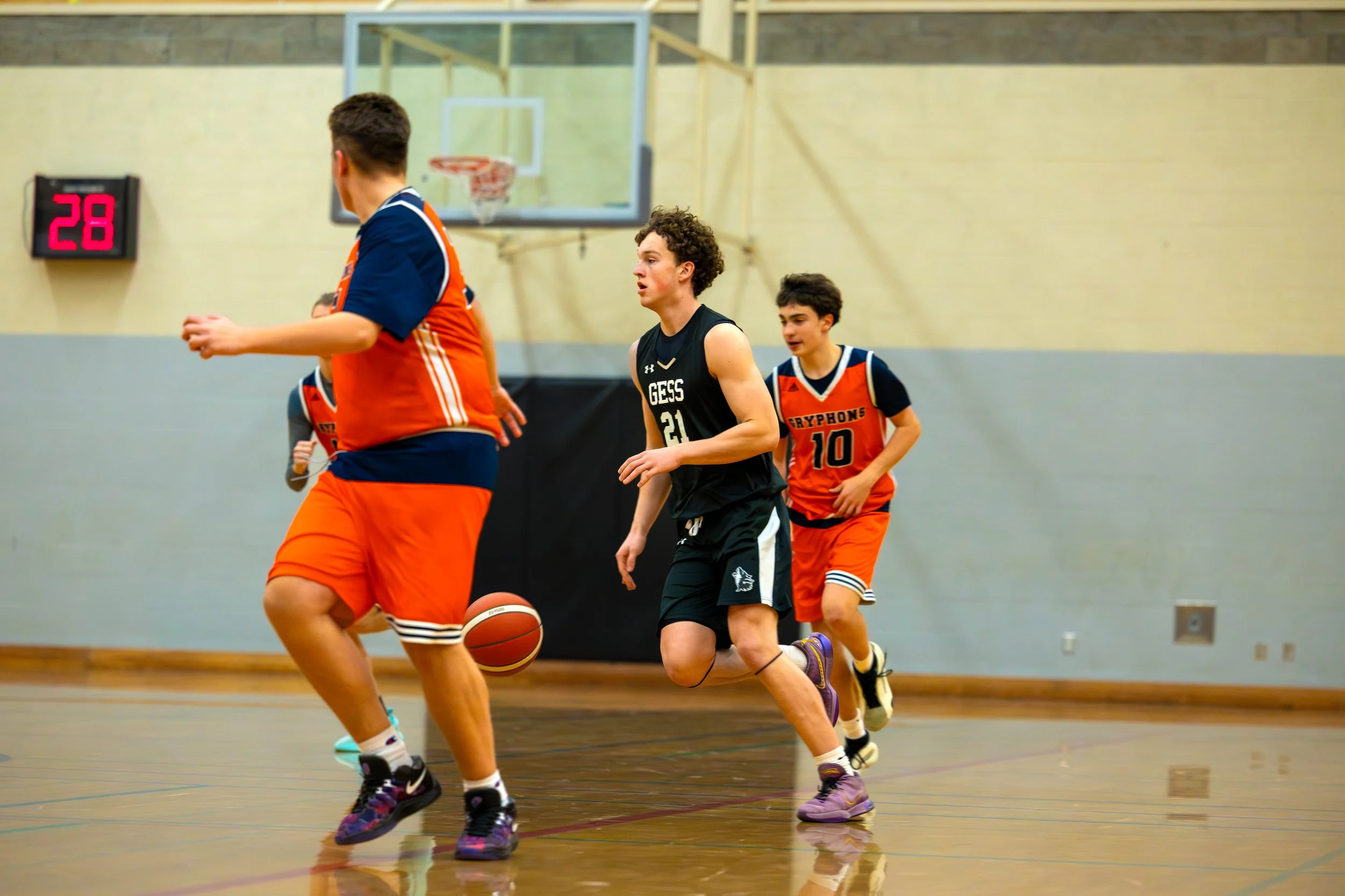 Young boys playing basketball indoors, with one in black uniform dribbling the ball and others in orange uniforms chasing.