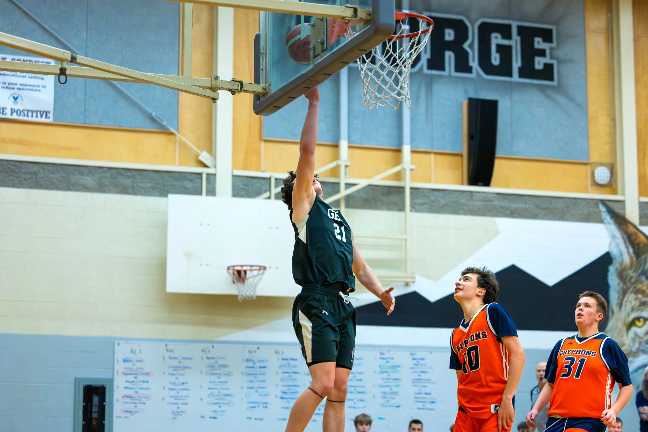 A youth basketball player in black jersey numbers 21 making a layup, with two players in orange and blue jerseys numbered 10 and 31 watching, in an indoor gymnasium.