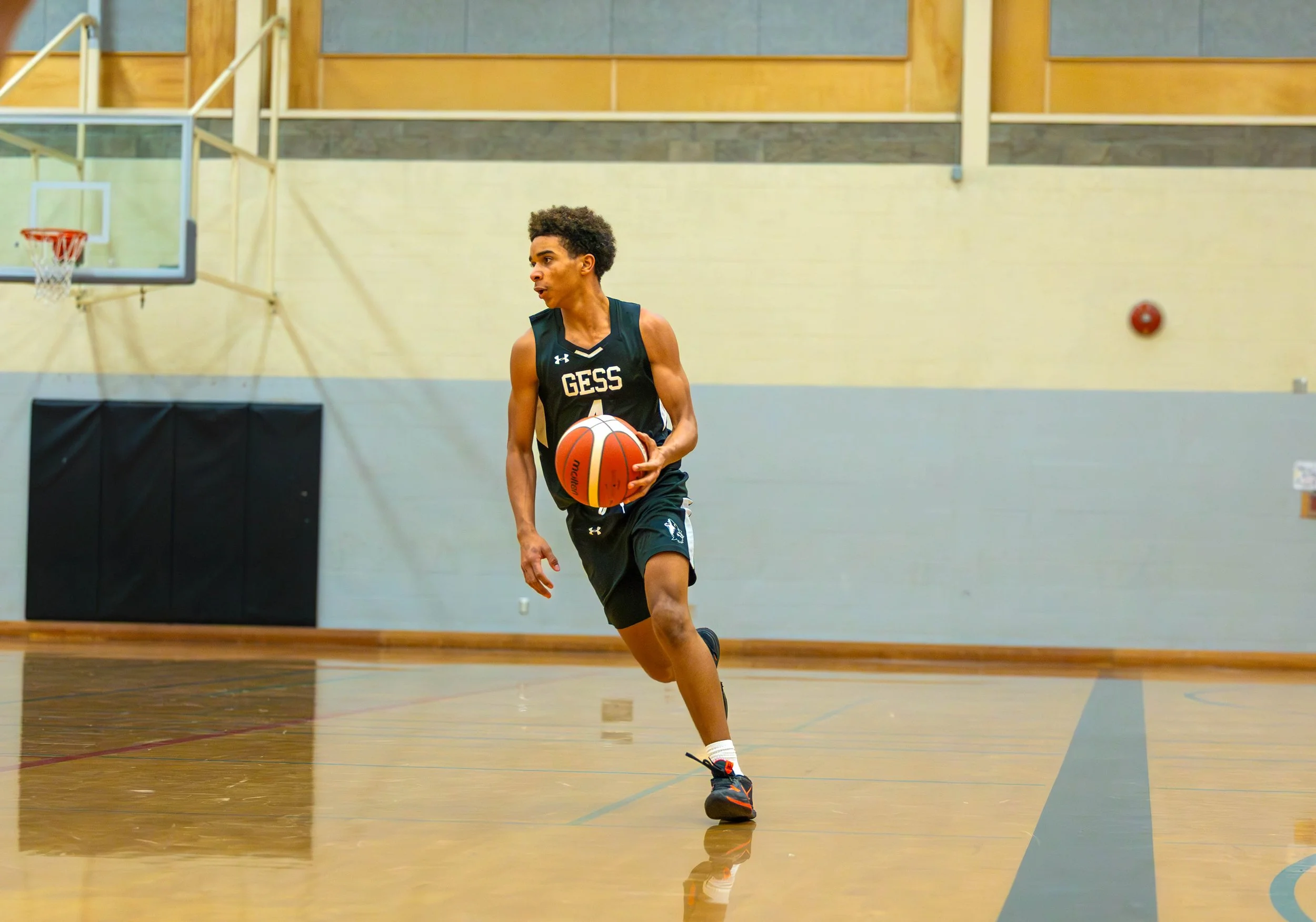 Young male basketball player dribbling on indoor court, wearing black uniform with 'GESS' on it.