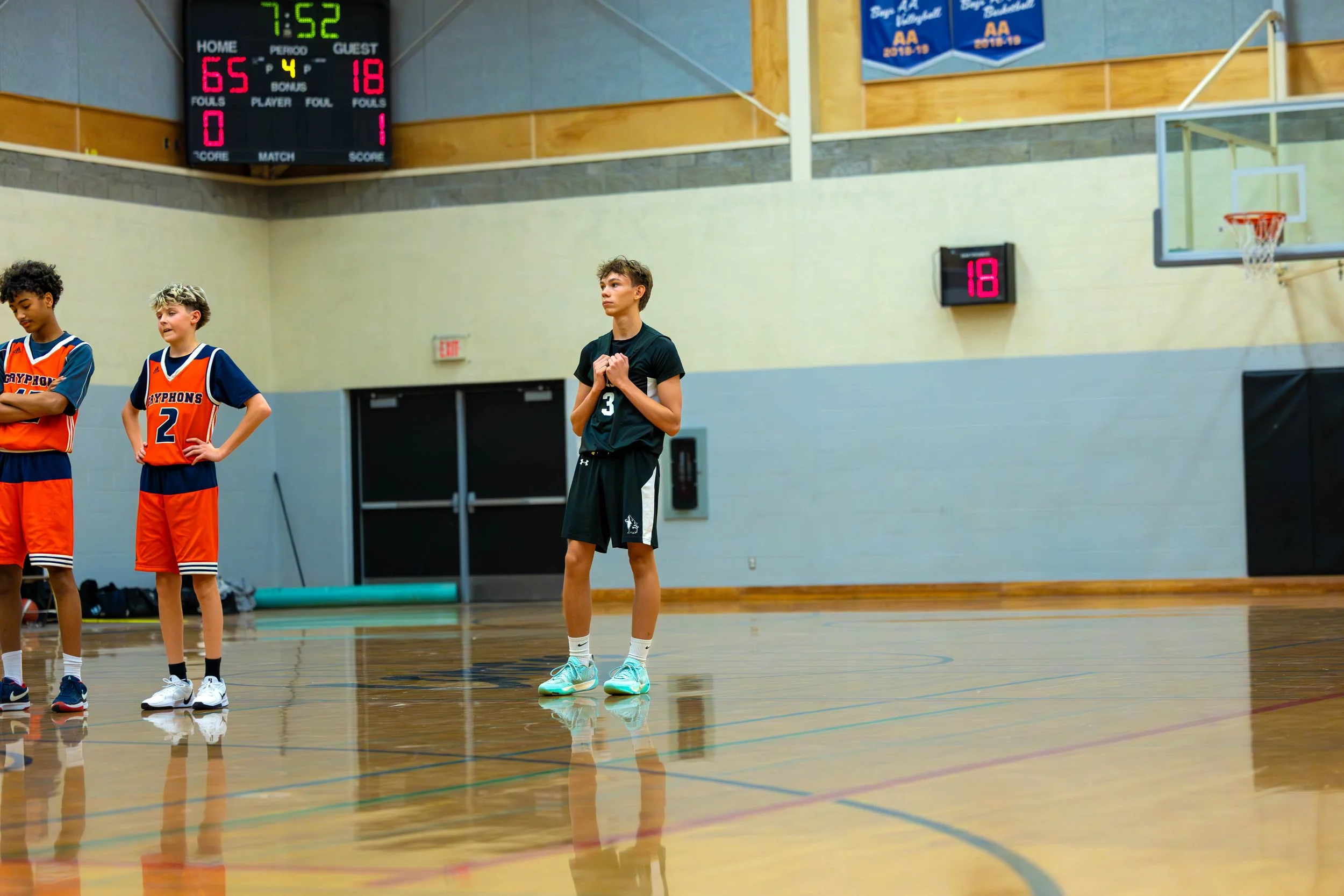 Young basketball players standing on a gym court during a game, with a scoreboard showing 7 minutes and 52 seconds remaining, and a score of 65 to 18.