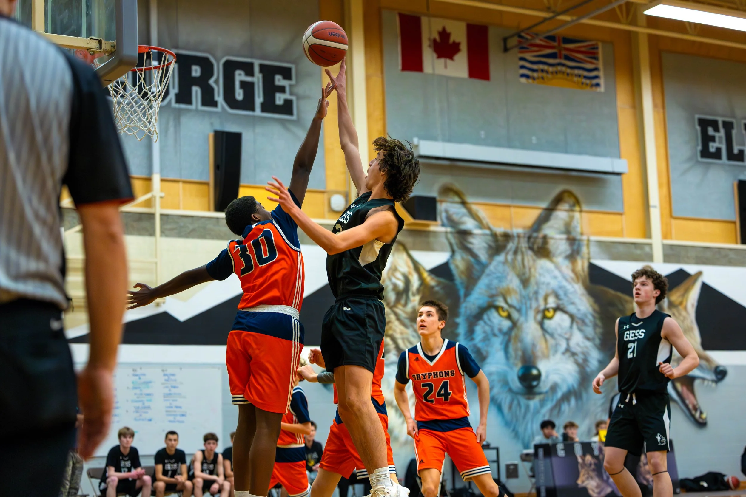 Young basketball players in a gym during a game, with one player jumping to shoot or block the ball, others watching or preparing, and a referee visible on the left. The gym has a mural of a wolf on the wall and flags hanging from the ceiling.