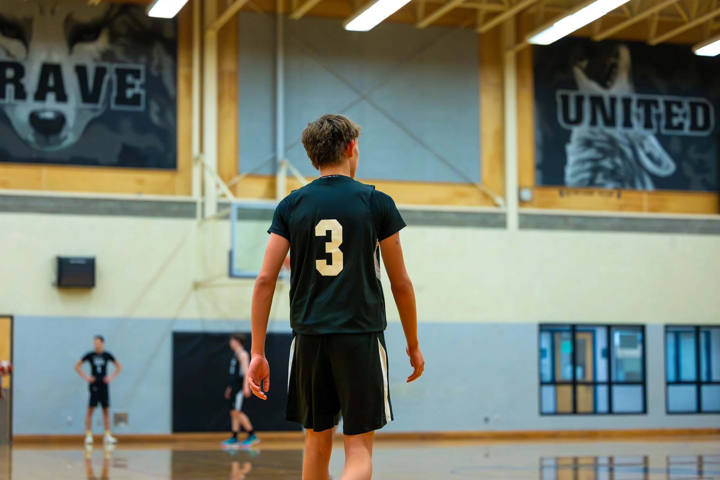 A young male volleyball player in a black uniform with the number 3 on the back, standing on an indoor court, facing away, with other players in the background, inside a gym with large banners on the wall.