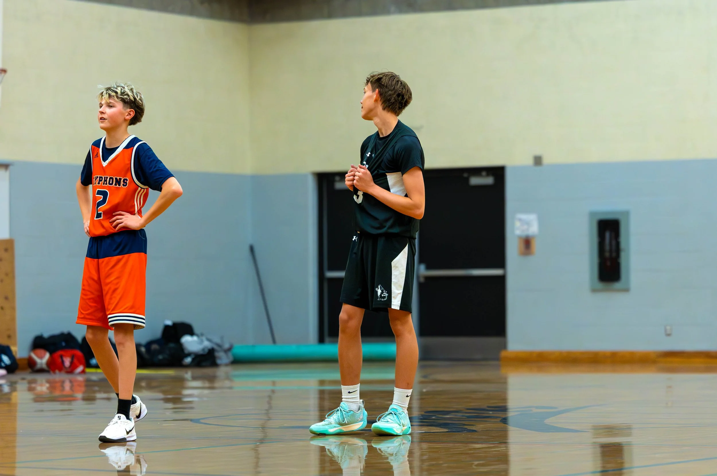 Two boys on an indoor basketball court, one in an orange and blue uniform with the number 2, and the other in black sportswear with a whistle, standing and talking.