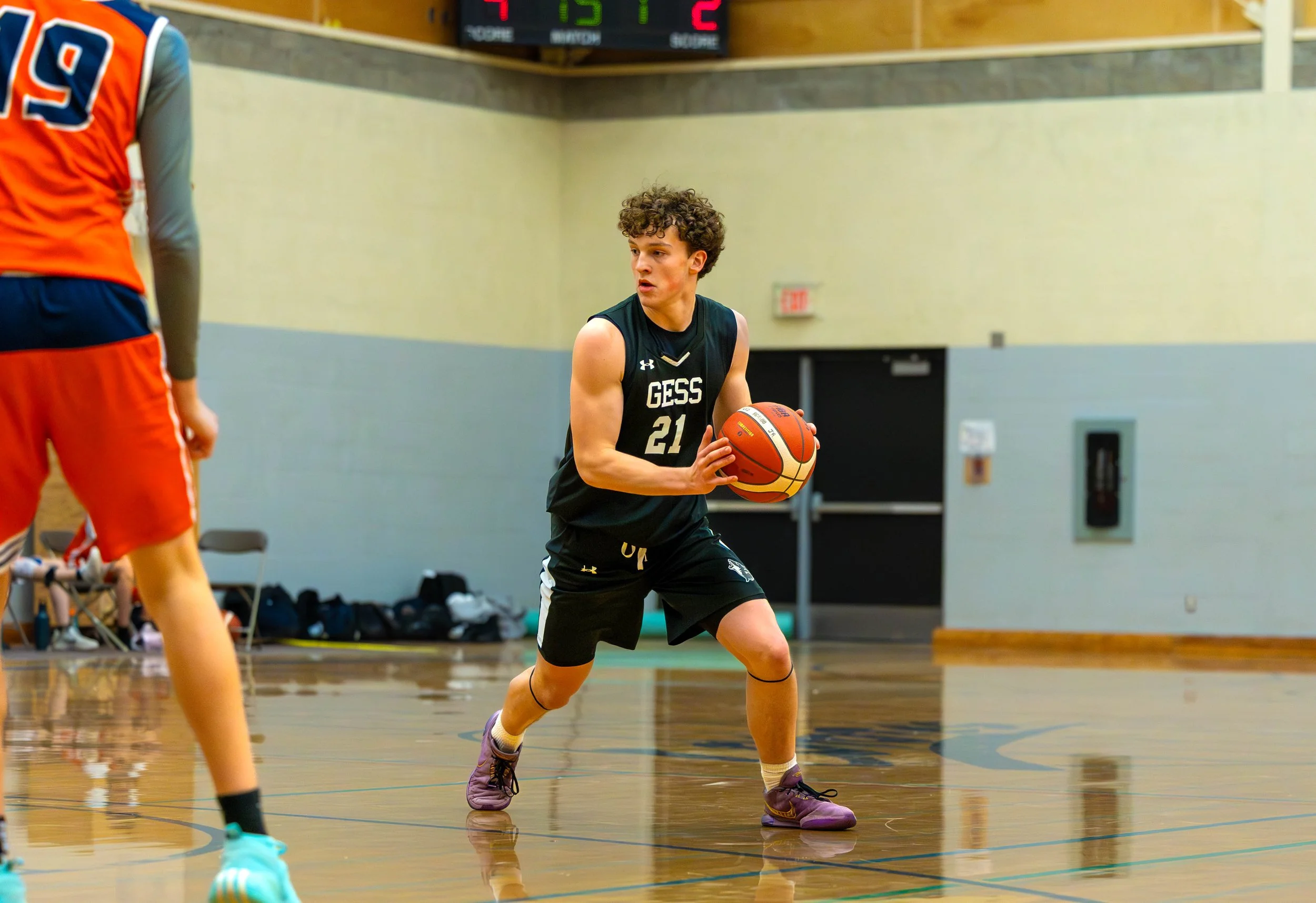 A young male basketball player wearing a black jersey with the word 'GESS' and the number 21, holding a basketball on an indoor court during a game.