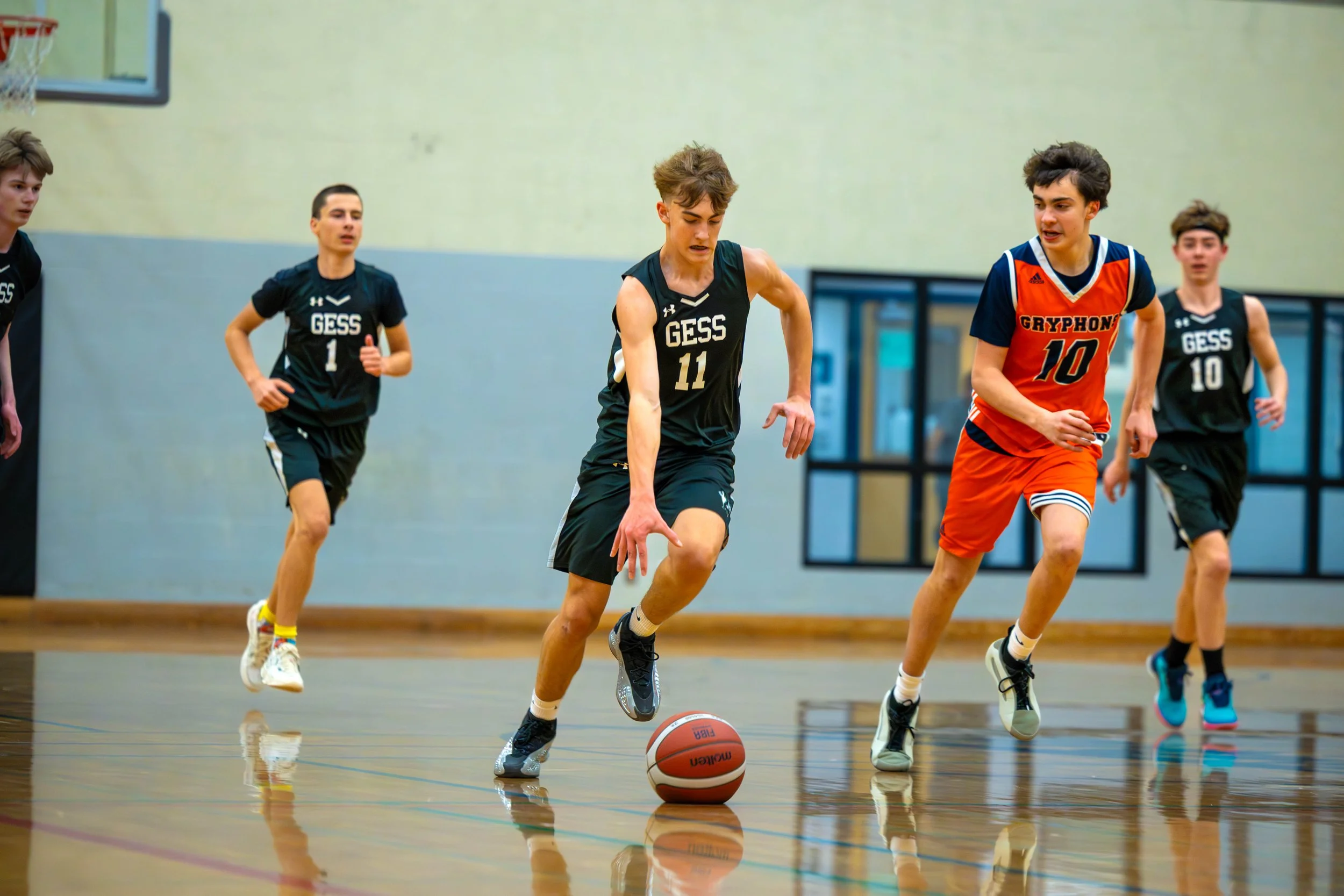 Young boys playing basketball indoors, two in black uniforms from GESS and one in an orange uniform from Gryphon, chasing after a basketball.