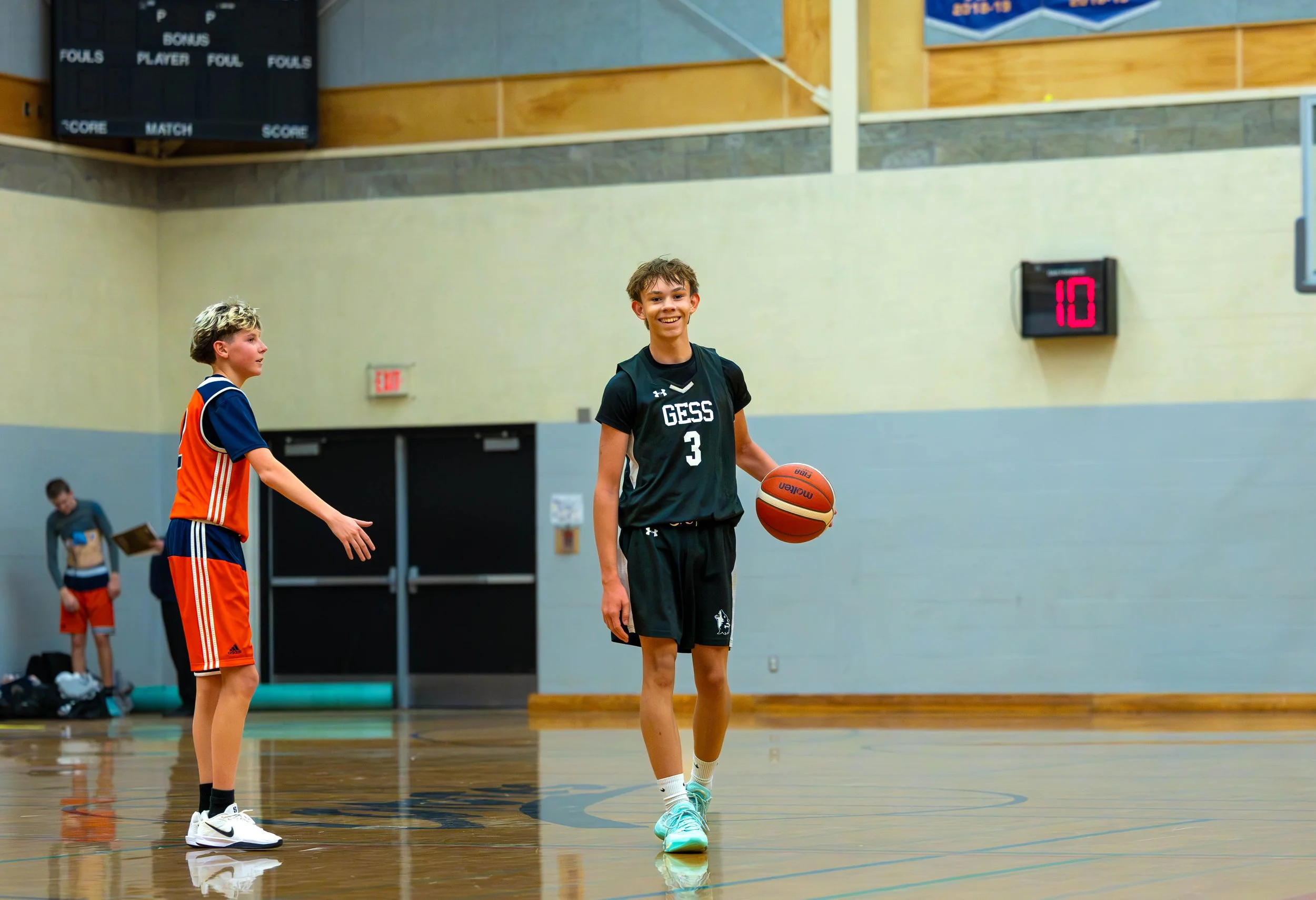 A young boy in a black basketball jersey with the number 3 and 'GESS' on it, holding a basketball, standing on an indoor basketball court. Another boy in an orange sports uniform is reaching towards him, and two more children are in the background. A