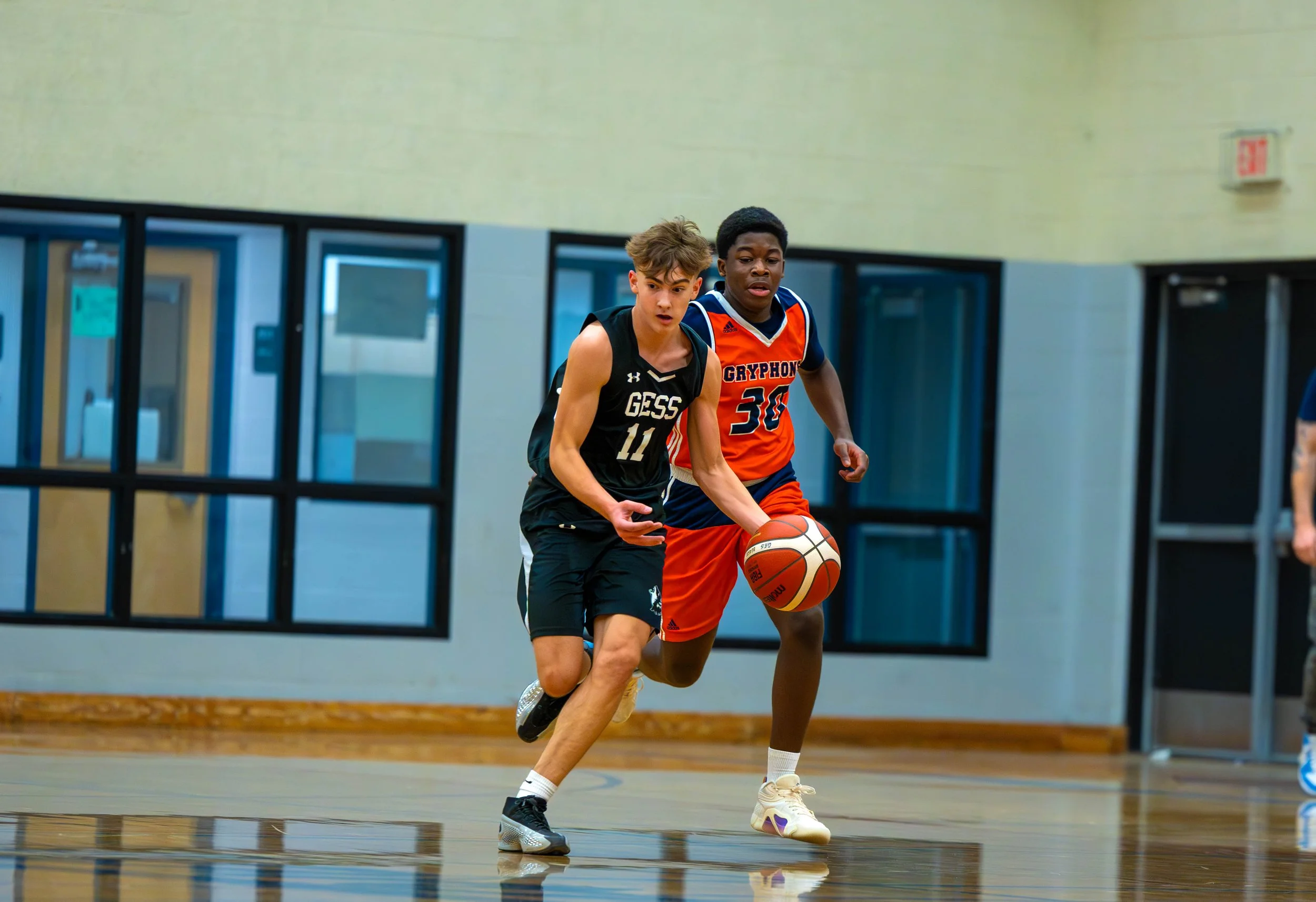 Two boys playing basketball in a gym, one in a black jersey with number 11 and the other in an orange jersey with number 30, running after the ball.